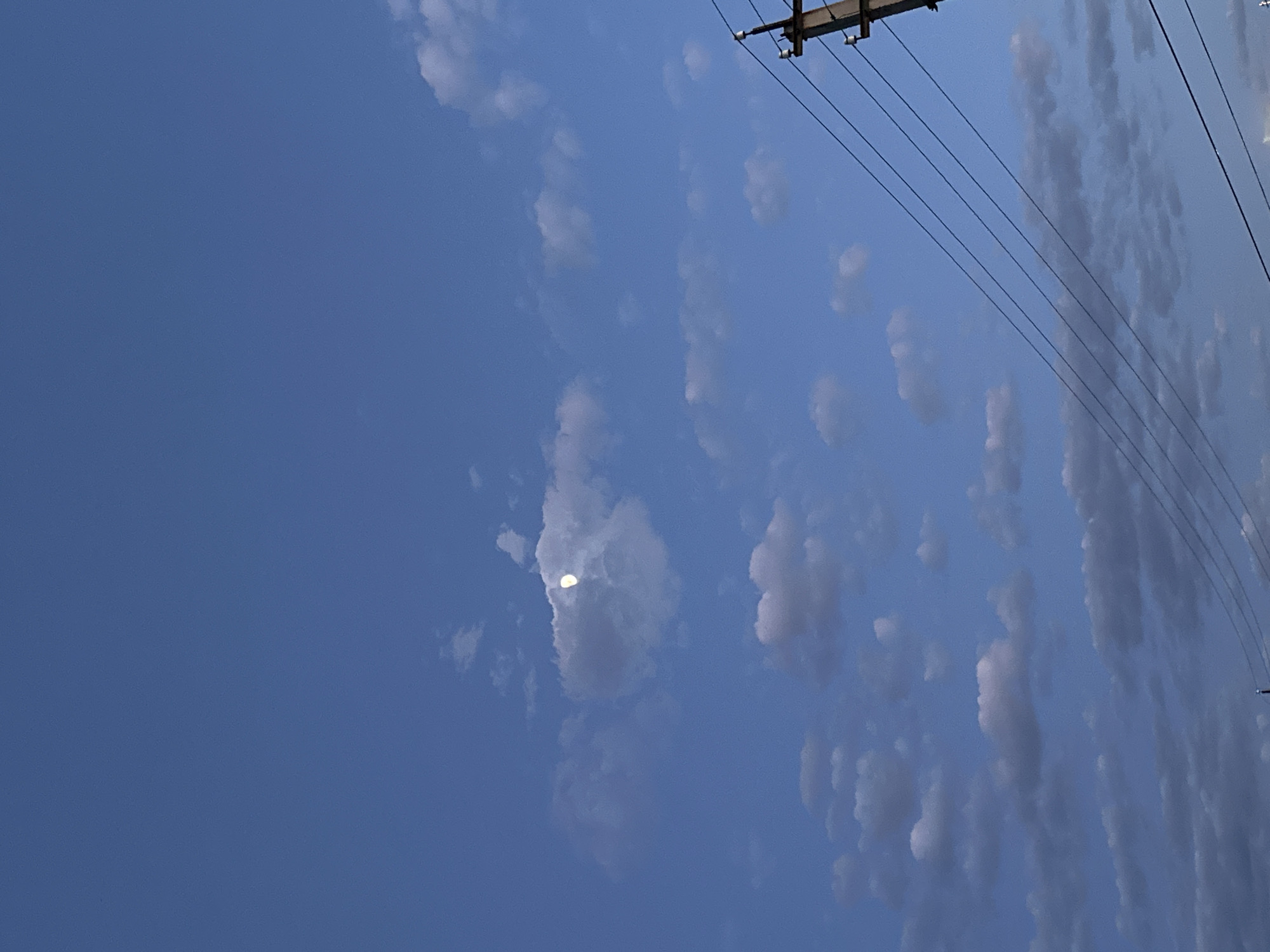 The image depicts a daytime sky with blue hues and scattered clouds, likely in Unknown, Unknown. Power lines and a utility pole are visible in the upper right corner of the frame. A partial view of the moon can be seen through a break in the clouds on the left side.