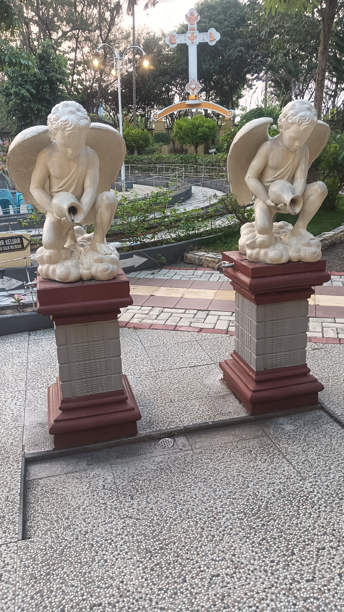 The image depicts a stone fountain sculpture of two cherubs holding pitchers, situated outdoors in what appears to be a garden or courtyard in Dukuh, Indonesia. The cherubs are depicted in a kneeling position on fluffy cloud-like bases, with water flowing from their pitchers. Each sculpture is mounted on a two-tiered pedestal with decorative brickwork. In the background, a white Christian cross with orange accents is visible, along with streetlights and lush green trees. The ground is paved with a combination of small pebbles and patterned tiles. A sign with Indonesian text is partially visible on the left side of the image, indicating an exit. The overall scene suggests a religious or memorial site.