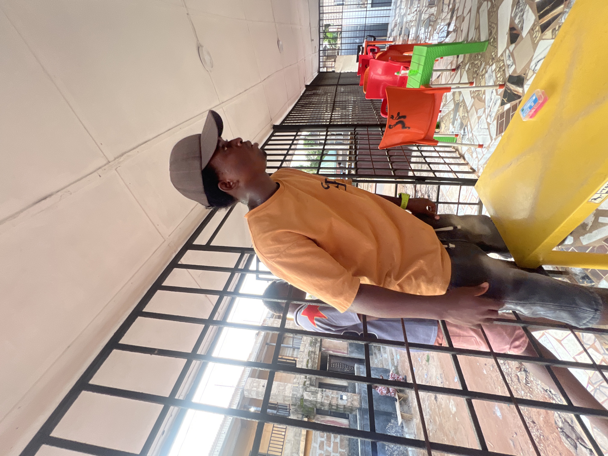 In Nkpor, Nigeria, a young Black man wearing an orange t-shirt and a gray baseball cap looks upwards while standing behind a black metal fence. The man's right hand is visible, resting on the fence. Behind him, through the fence, several colorful plastic chairs are arranged. A bright yellow structure, possibly a table or bench, is on the right side of the image. The flooring in the background is tiled with a mosaic pattern. Further in the background, a building with multiple windows and balconies can be seen. The scene appears to be outdoors, possibly in a public space or a seating area. The lighting suggests it is daytime.