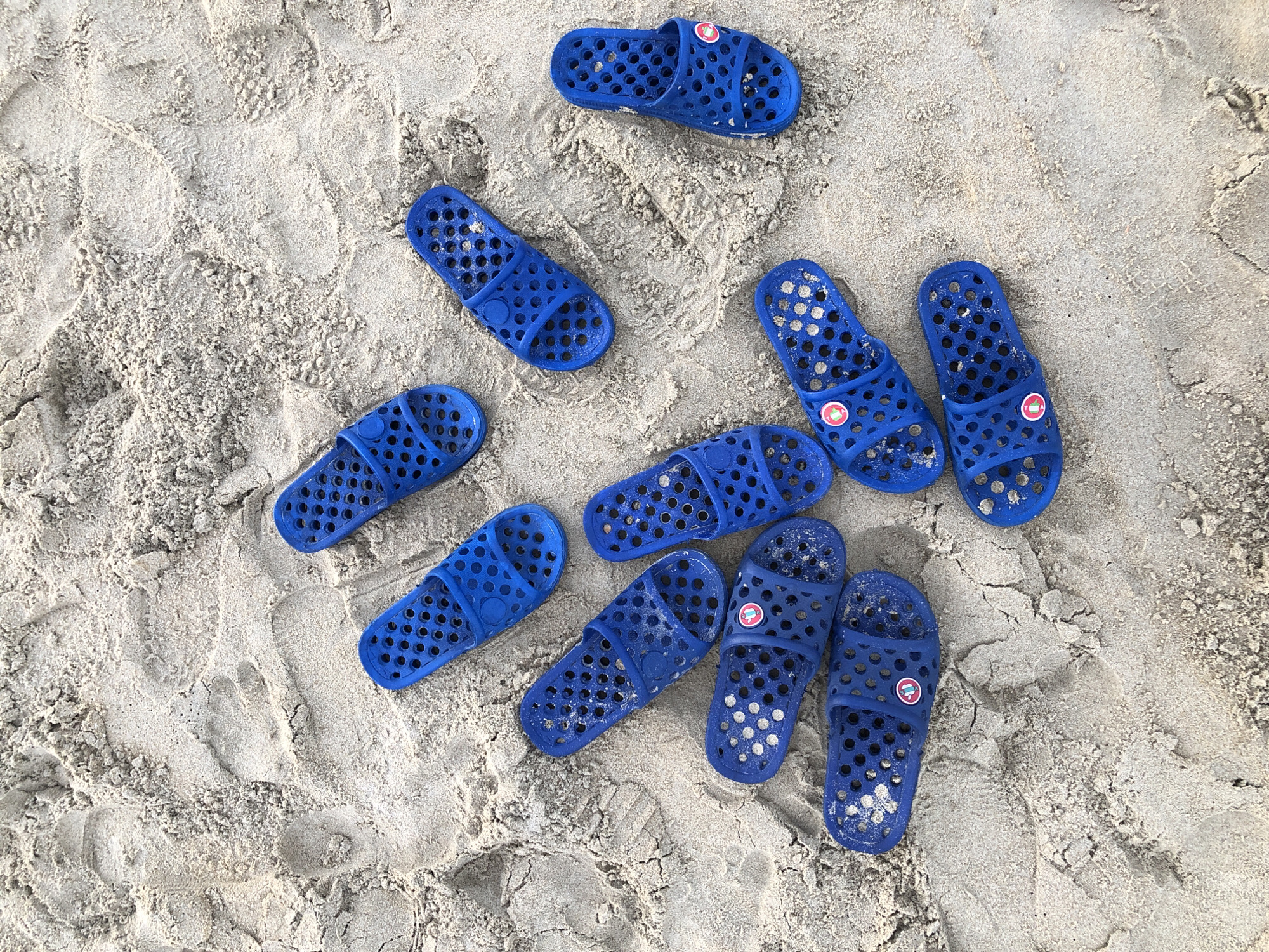 The image shows a collection of ten blue rubber slide sandals scattered on a sandy surface. The sandals are all of a similar design, featuring a single strap with multiple circular holes for drainage. Several of the sandals have a small circular logo with a red and white design on the strap. The sand appears slightly damp in some areas, suggesting it might be near water. The lighting suggests it is daytime, possibly a sunny day given the bright white of the sand. The scene is set outdoors on a beach, likely in a tropical or warm climate. The presence of multiple sandals implies they belong to a group of people, possibly a family or friends enjoying a day out. There are some faint footprints visible in the sand, indicating recent human activity. There is no visible text in the image. The overall impression is one of leisure and relaxation in a coastal environment. The context provided is that this scene is located in Đà Nẵng, Vietnam, which is known for its beaches.