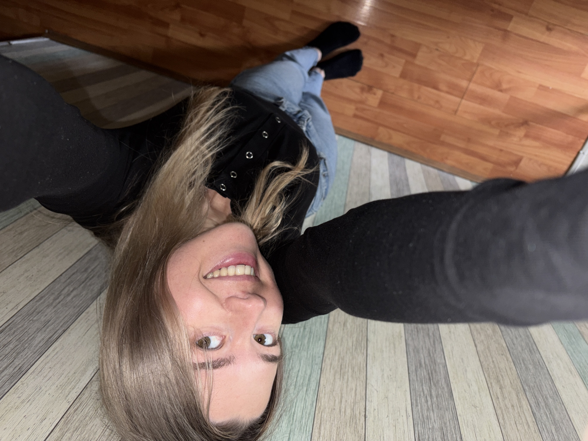 A young woman is captured in a low-angle selfie, lying on her back on an indoor floor. She appears to be in her 20s or 30s, with light brown or blonde hair falling around her head. She is looking directly up at the camera with a wide, cheerful smile, her eyes sparkling with amusement. Her arms are extended towards the camera, likely holding the device.
She is dressed in a black long-sleeved top, possibly a fitted top or a corset-style garment, featuring a line of silver eyelets or buttons down the front. She is also wearing light blue jeans and black socks. Her legs are visible in the background, crossed at the ankles.
The setting is indoors, featuring a distinctive floor composed of two different types of planks. One section, primarily in the upper right, is a warm, light brown wooden or laminate flooring. The other section, covering the left and lower parts of the image, consists of striped planks in shades of grey, white, and light blue. The transition between these two flooring types creates a visual division in the scene. The lighting suggests an indoor environment, possibly with artificial light, given the absence of strong natural light sources. The overall mood conveyed by the woman's expression is one of happiness and playfulness. The location is contextualized as Chaikovsky, Russia. There is no visible text in the image.