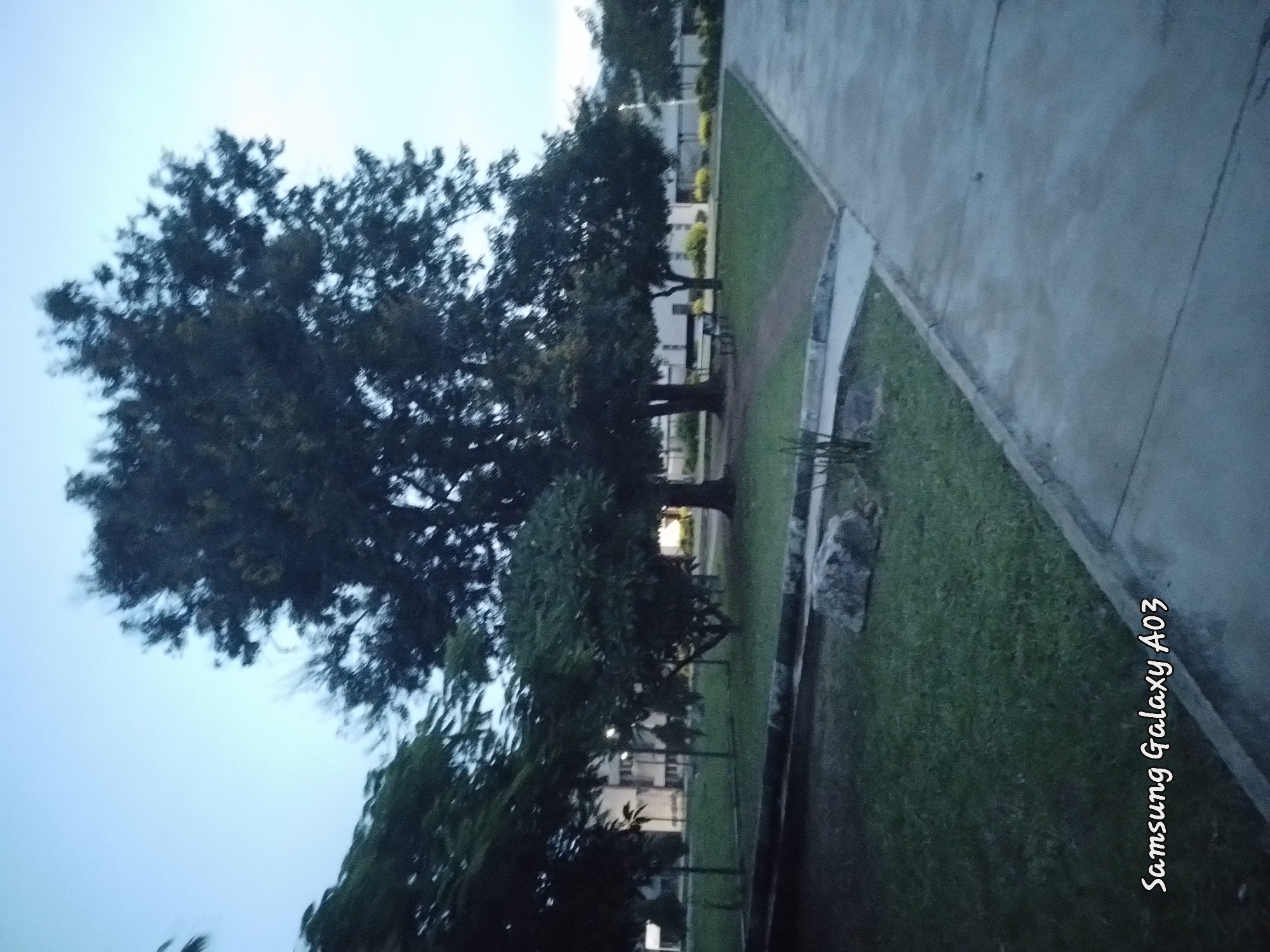 This is a low-light photograph taken outdoors, possibly at dusk or dawn. The scene depicts a paved walkway running alongside a grassy area with several trees. In the background, there are low-rise buildings. The photograph also includes a watermark in the bottom left corner that reads "Samsung Galaxy A03". The location of Jos, Nigeria is not explicitly indicated in the image itself, and therefore cannot be confirmed.