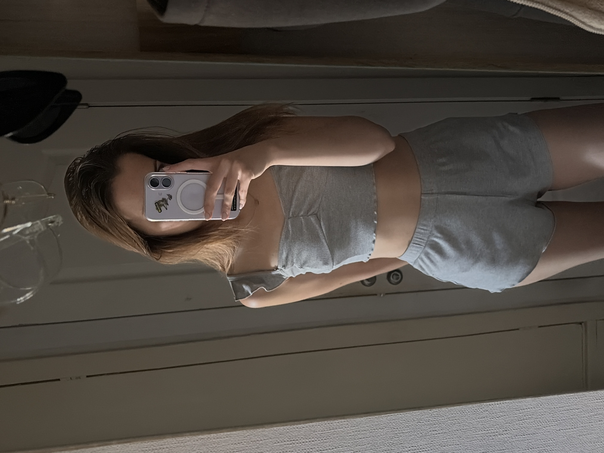 The image is a full-body mirror selfie taken by a young woman with long, light brown hair. She is wearing a grey ruched crop top with ruffled straps and matching grey shorts. She is holding a light purple smartphone in her right hand, obscuring her face. The background appears to be an indoor setting, possibly a bedroom or dressing area, with a white paneled door and a textured wall to the left. There are reflections in the mirror of what appear to be a light fixture and a dark object, possibly a hat or lamp, in the background. The overall lighting is dim. There is no visible indication of the location being in Kyiv, Ukraine.
