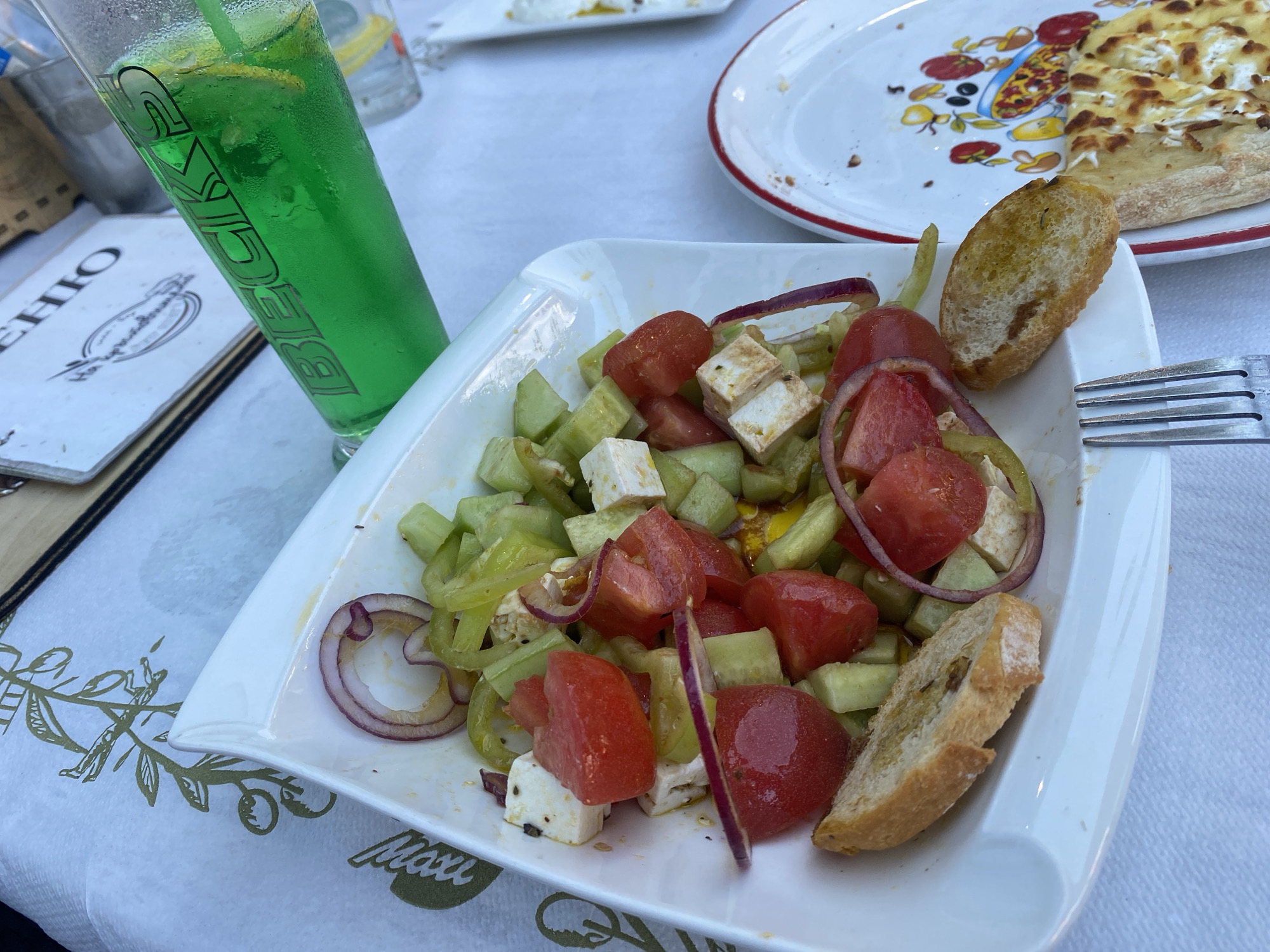 The image depicts a dining scene at Pizzeria & Grill Maxi in Smolyan, Bulgaria, identifiable by the "Maxi" branding in an olive branch motif on the white paper tablecloth. A meal is set on a light-colored table, likely outdoors or in a brightly lit indoor space.
In the foreground and center, a rectangular white dish holds a Greek-style salad. The salad consists of roughly diced red tomatoes, chopped green cucumbers, cubes of white feta cheese, and thin slices of red onion. A thin layer of olive oil is visible at the bottom of the plate. Two slices of toasted bread, possibly garlic bread or bruschetta, are positioned on the edge of the salad plate, one standing upright and the other lying flat. To the immediate right of the salad, a silver four-pronged fork rests on the tablecloth.
To the left of the salad, a tall, frosted glass contains a vibrant green beverage with ice and a visible lemon slice. The glass has the word "BECKS" printed vertically in dark lettering. Partially visible beneath the glass is an open menu or booklet with a light-colored cover and dark text, including what appears to be "EHIO" at the top.
In the upper right background, a round white plate with a distinctive red rim holds a partially consumed baked dish. This dish appears to be a flatbread or pizza base topped with melted white cheese, with some areas golden brown from baking. The plate itself is decorated with colorful illustrations of food items, such as olives and peppers, around its edge.
The overall scene indicates a casual dining experience, with no individuals visible in the frame.