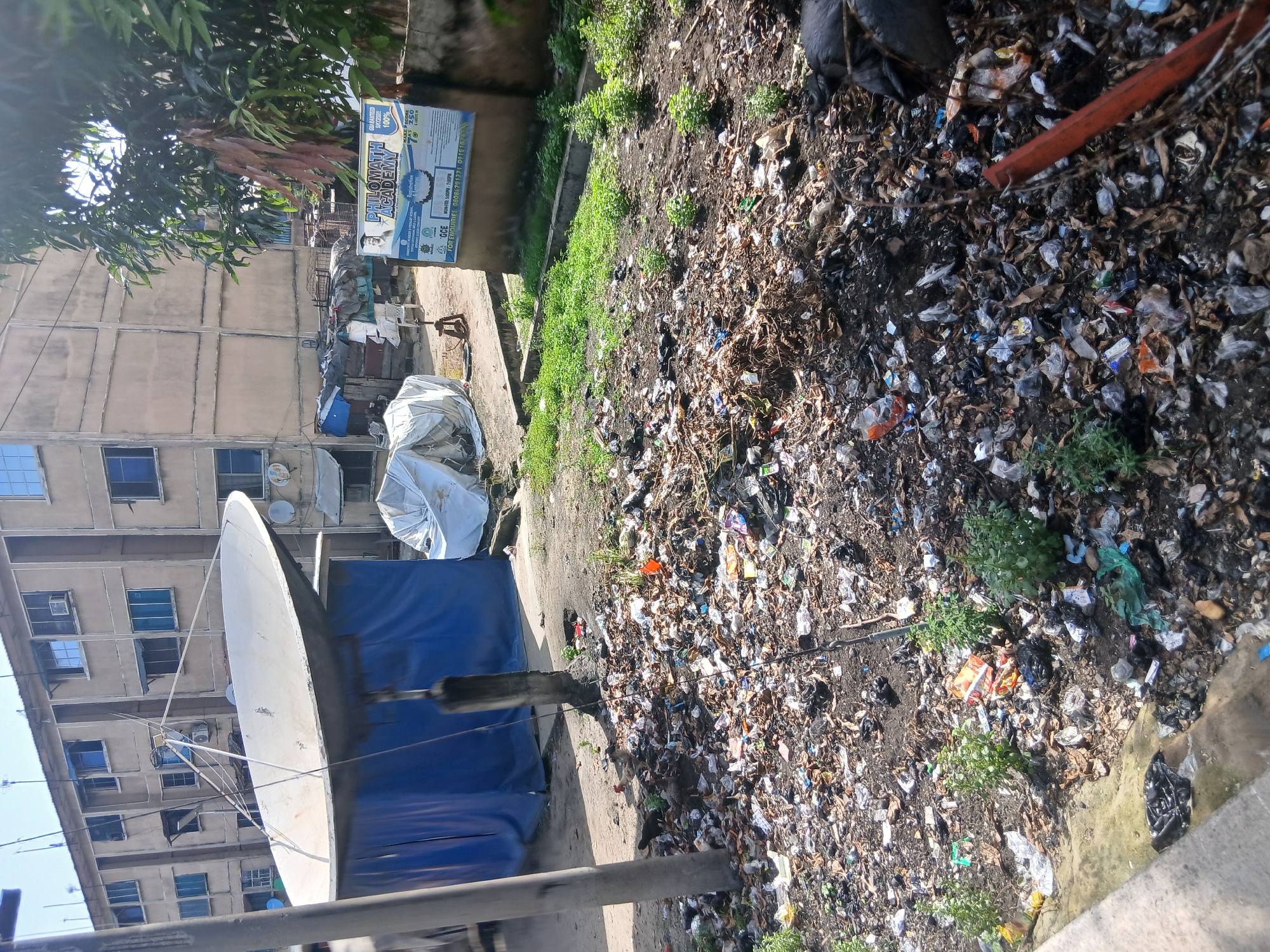 The image depicts a scene in Lagos, Nigeria. A large amount of litter and refuse covers the ground. A satellite dish stands in the foreground. In the background, a multi-story building with multiple windows is visible. A sign for "PHILOMATH ACADEMY" is attached to a wall. Bags of trash are present.