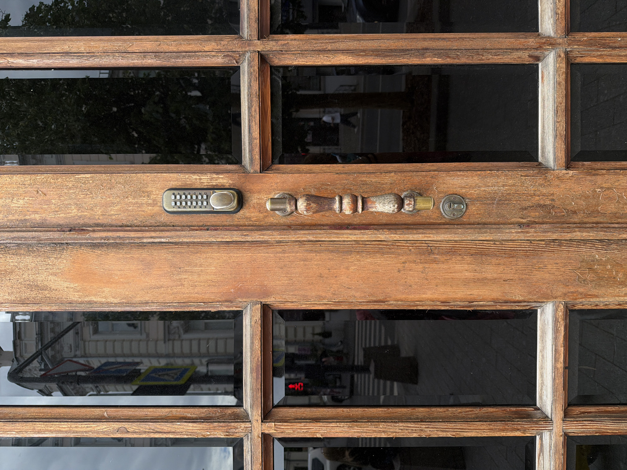 The image depicts a close-up view of a weathered wooden door with multiple glass panels. The door features a keypad, a handle, and a keyhole. The glass panels reflect the surrounding urban environment, including trees, buildings, and street signs. A red LED arrow sign is visible through one of the panels. The overall location is an urban area, specifically in the city of Unknown, Unknown.
