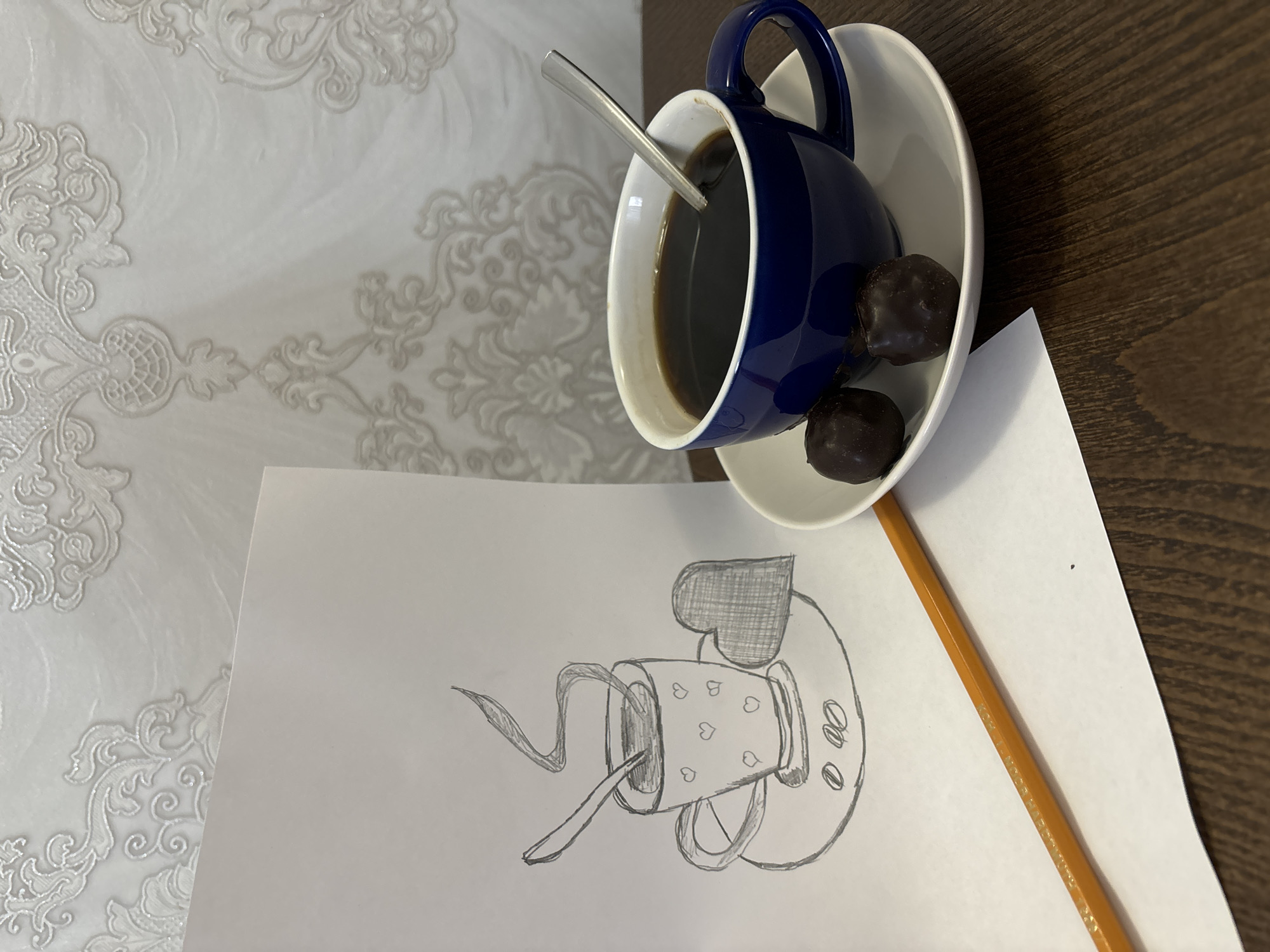 The scene, located in Chaikovsky, Russia, presents an indoor still life composition. A dark brown wooden surface forms the base of the image. On this surface, a white sheet of paper is positioned on the left, displaying a monochrome pencil drawing. The drawing features a stylized steaming cup with heart patterns, containing a spoon, next to a shaded heart shape, all resting on a saucer with two oval shapes resembling coffee beans. An orange wooden pencil lies diagonally across the lower portion of the paper, partially obscuring text on its side. To the right, a dark blue ceramic cup, filled with a dark liquid and containing a silver-colored spoon, rests on a white saucer. Two dark chocolate truffle-like candies are placed on the saucer beside the cup. The background is a light-toned wall covered in wallpaper adorned with a shimmering, ornate, classical scrollwork pattern in a pale silver or grey hue. No human subjects are visible.