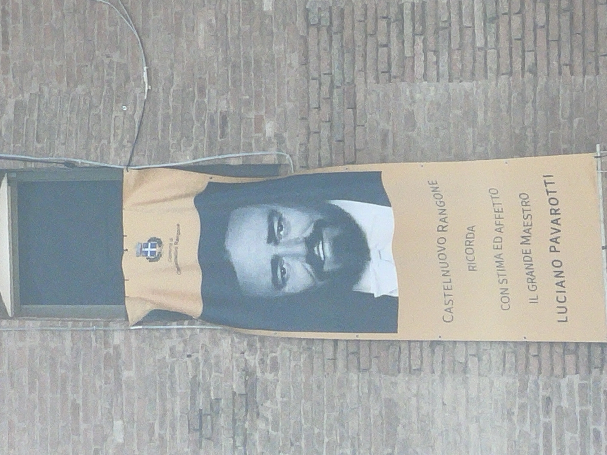 A banner hangs on a brick wall, featuring a portrait of Luciano Pavarotti and text. The banner is yellow with a dark blue image of Pavarotti's face, a white bow tie, and a dark blue jacket. The text on the banner reads "CASTELNUOVO RANGONE RICORDA CON STIMA ED AFFETTO IL GRANDE MAESTRO LUCIANO PAVAROTTI". Above the portrait, a coat of arms is visible, along with the words "Comune di Castelnuovo Rangone". The banner is suspended from above by wires and poles. The brick wall is made of light brown bricks and shows signs of age. A window is visible behind the upper part of the banner. The scene is outdoors, suggesting a public space in Castelnuovo Rangone, Italy.