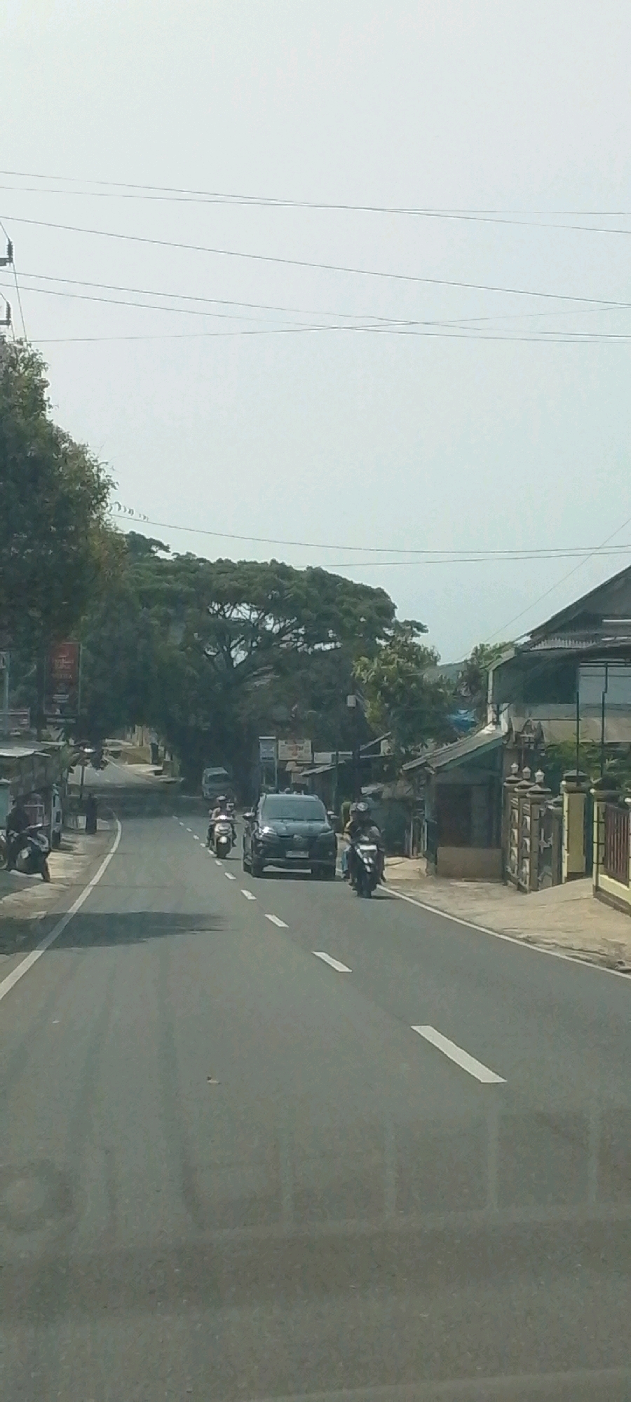 The image depicts a two-lane asphalt road in Susukan, Indonesia, under a hazy, overcast sky. The road has white dashed lines marking the lanes and solid white lines on the outer edges. On the left side of the road, there are parked motorcycles and what appear to be small roadside businesses with signage. Lush green trees and vegetation line the background, with some buildings visible on the right side of the road.
Several vehicles are present on the road. In the foreground, a black SUV is driving in the right lane, followed by a motorcycle. Further down the road, there are two more motorcycles, one in each lane, and another vehicle further in the distance. The general atmosphere suggests a moderately busy, semi-urban or rural environment.
