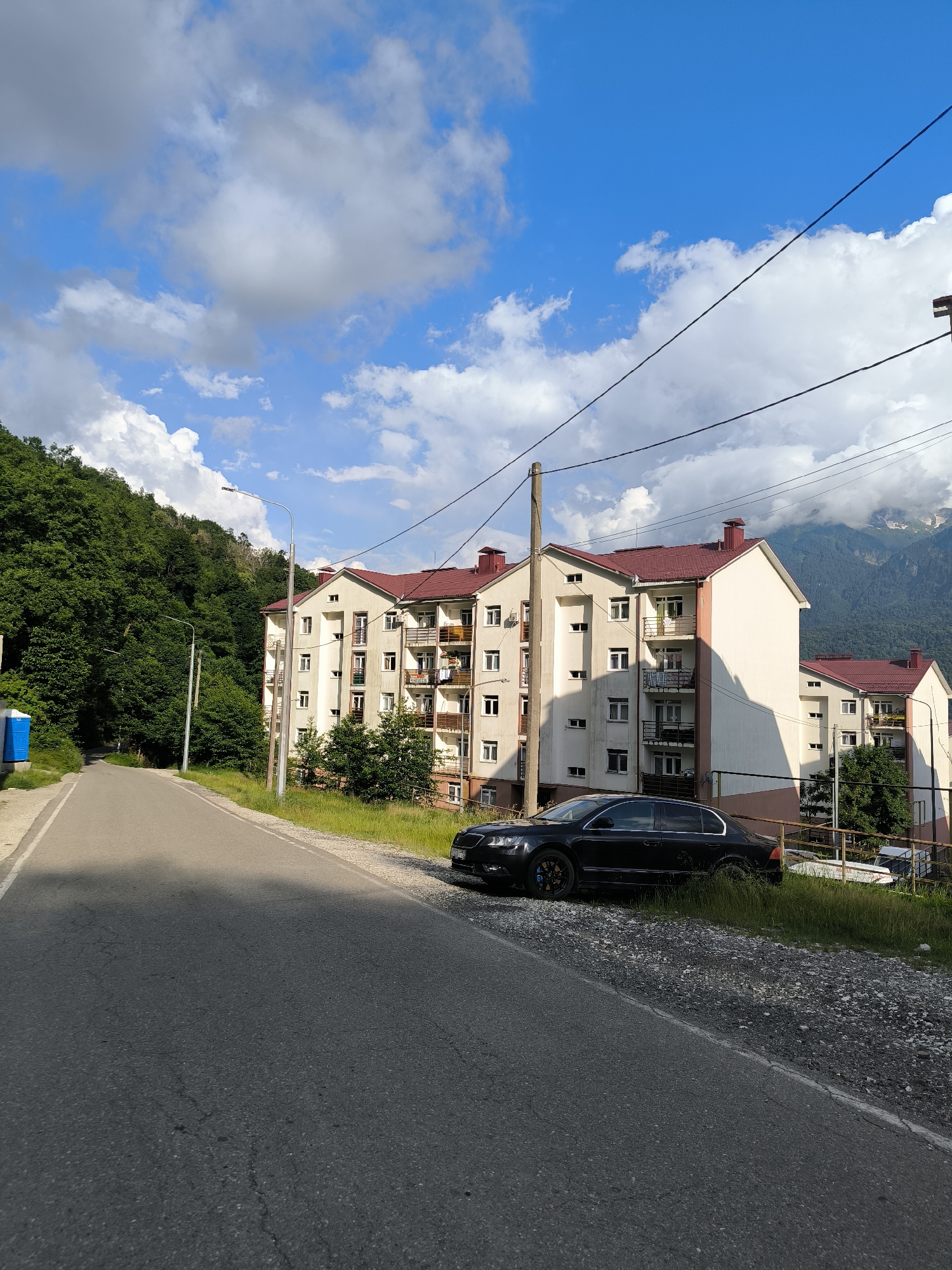 This media file depicts an outdoor scene in Krasnaya Polyana, Russia, on a sunny day with scattered clouds. The focal point is a multi-story residential building with a reddish-brown roof, white and pink siding, and balconies. A black sedan is parked on the side of an asphalt road in front of the building.
To the left of the building, a lush green, tree-covered mountainside slopes upwards, suggesting a mountainous or hilly terrain. A blue portable toilet is visible on the side of the road on the left. Streetlights are present along the road. In the background, distant mountains are visible, some with snow-capped peaks, indicating a higher altitude. The sky is a vibrant blue with fluffy white clouds. The lighting suggests it is daytime, likely late morning or afternoon. No people are visible in the image. The overall mood is peaceful and serene, characteristic of a natural, mountainous landscape combined with a residential area.