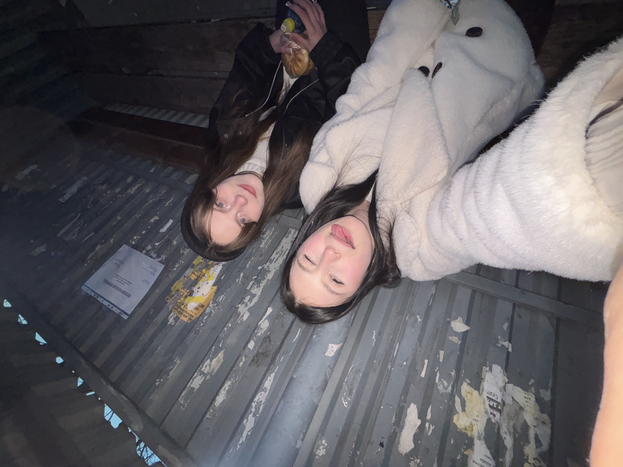Two young women lie on a weathered, corrugated metal surface littered with torn paper and debris. One woman, with light brown hair, wears a black jacket over a white turtleneck and holds a beverage and wrapped food. The other woman, with dark hair, wears a fluffy, light-colored jacket. The photo is taken from a low angle, looking up at the women. The location is Фоки, Russia, as specified. The background shows glimpses of the sky through gaps in the metal.