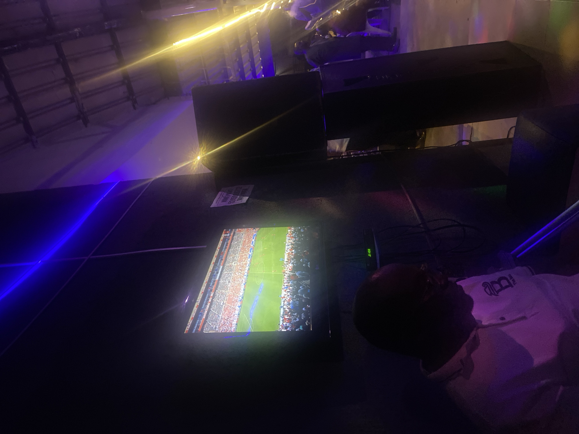 The scene depicts an indoor entertainment venue in Jos, Nigeria, characterized by dim lighting and prominent colored illumination. A large, dark flat-screen television dominates the center-left, displaying a soccer (football) match on a green field with players and a crowded stadium visible. To the right of the TV, two large, dark speakers are stacked vertically. A white sign partially affixed to the wall next to the speakers reads "MORE INFO" and includes a phone number: "53450309712". Below the television, a piece of equipment displays a small green digital readout of ".0100".
In the foreground, a dark-skinned man with a balding head and glasses is seated, facing away from the camera and towards the screen. He wears a light-colored polo shirt featuring a dark, stylized "B" logo and the text "The Best" on the left chest. His face is largely obscured by shadow.
The environment is illuminated by distinct light sources: a strong blue laser or light beam crosses the upper left portion of the image, and a bright yellow light source emanates from the upper right, casting significant glare and light streaks across the scene. The ceiling above is an exposed industrial style with visible trusses and beams. In the right background, several individuals are present, some standing and others seated on white chairs, largely silhouetted by the yellow light and deep shadows. The floor appears to be concrete or a similar material, reflecting some of the colored lighting. Cables are visible behind the television and speakers, indicating electronic setup.