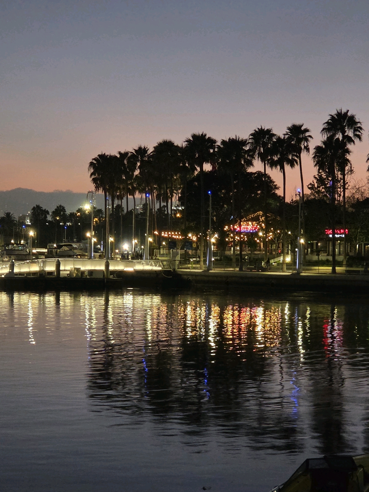 This is a nighttime photograph taken at Shoreline Village in Long Beach, California. The image depicts a waterfront scene with palm trees silhouetted against a dusky sky. Numerous lights from buildings and along the waterfront are reflected in the calm water of the harbor. A bridge with illuminated railings is visible in the middle ground, and several boats are docked in the marina. The overall atmosphere is serene and festive, with the colorful lights creating a warm glow.
