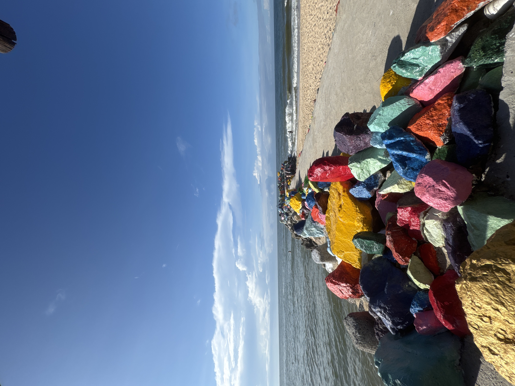 A concrete pier extends into the ocean, lined with vibrantly painted rocks in various colors. The pier is situated on a sandy beach with gentle waves lapping at the shore. The sky is blue with scattered white clouds. In the distance, a person can be seen swimming or wading in the water. The rocks are positioned on both sides of the pier, with a denser cluster on the left, closest to the viewer. The scene appears to be a public art installation or decorative feature along the coastline of Phan Thiết, Vietnam.