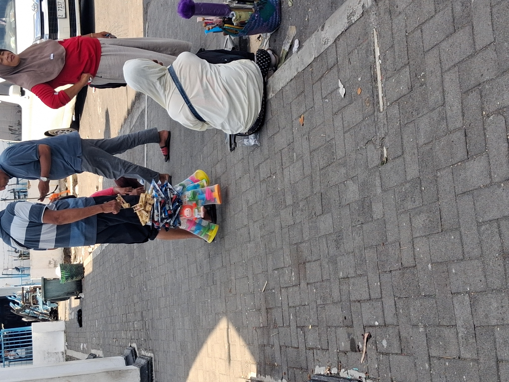 The image depicts an outdoor scene on a paved walkway in the city of Surakarta, Indonesia. In the foreground, a person, partially obscured from the torso down, stands holding a stack of small, colorful toys. The toys appear to be small figures or trinkets packaged for sale. To the right of this individual, another person is seated on the ground, wearing a light-colored hooded jacket and dark pants with white polka dots. This person appears to be interacting with a basket filled with various items, including what appear to be pens, brushes, and a purple, fuzzy object.
Further in the background, a woman wearing a red shirt and a brown hijab stands with her hand on her hip. She is looking towards the right of the frame. Next to her, a man in a blue and white striped shirt and dark shorts is walking away, holding a phone or similar device in his hand. Behind them, a white vehicle, possibly a van, is parked. Several green trash bins and some metal fencing are visible in the background, suggesting a public area or market setting. The ground is paved with interlocking rectangular bricks, and sunlight casts shadows across the scene.