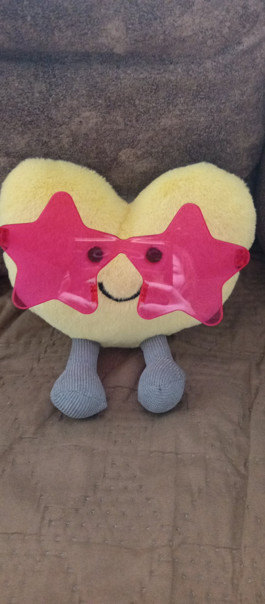 A plush, yellow, heart-shaped toy with grey knitted legs sits on a brown patterned surface. The toy wears large, pink star-shaped glasses and has a stitched-on smiling mouth and circular eye details behind the glasses. The toy is the primary focus of the image. The location is not discernible and is unknown.
