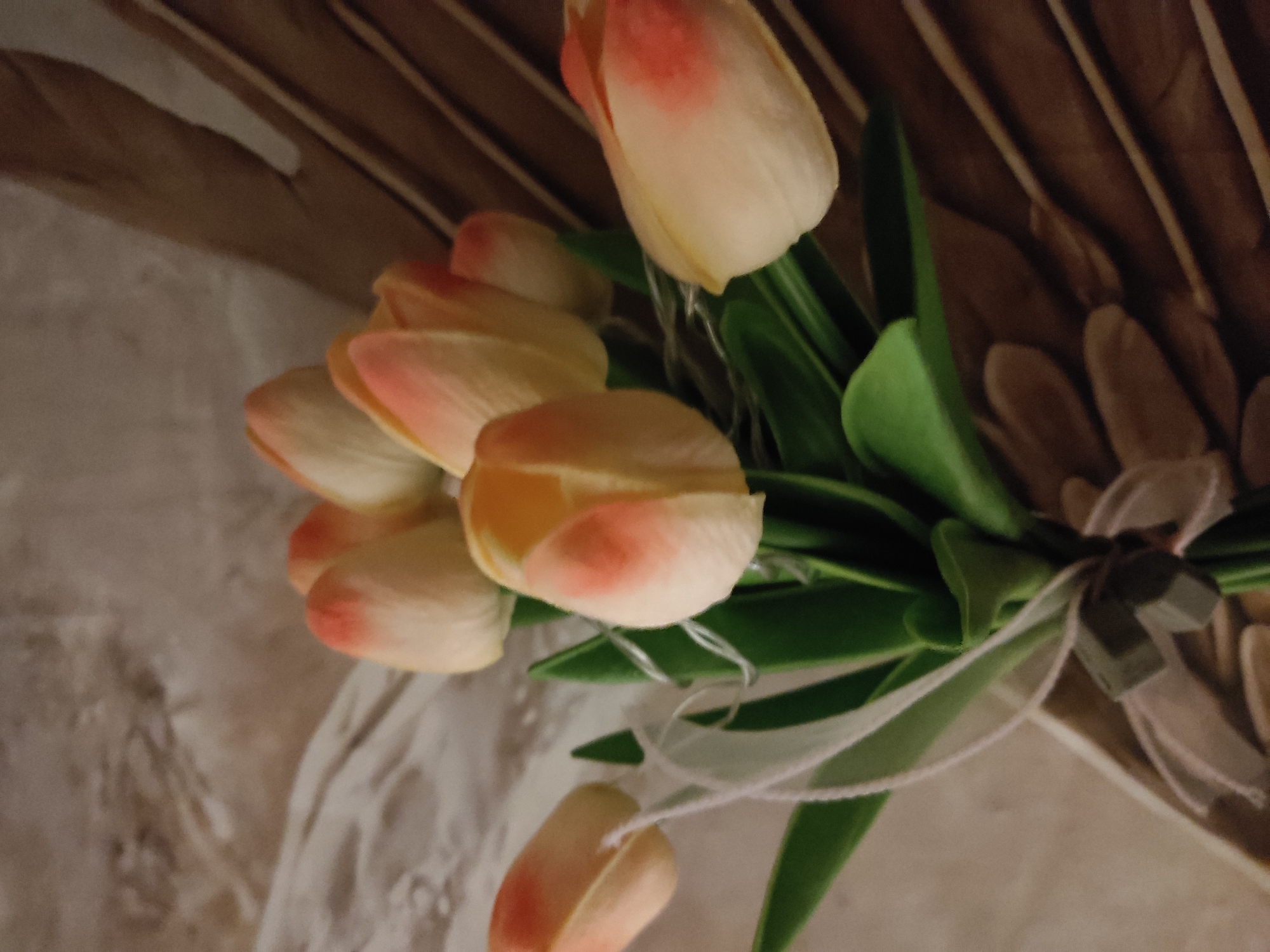 A bouquet of light pink and cream tulips with green leaves and a white ribbon is arranged in the foreground against a textured beige and brown background. The tulips have delicate pink tips and creamy white petals. The arrangement is tied with a white ribbon that has a small, dark, geometric ornament. The background features abstract patterns, with the left side showing a lighter, mottled texture and the right side displaying vertical brown stripes. This image was taken indoors, likely in a home or studio setting, with artificial lighting. There are no people or animals visible in the image. The location in Noginsk, Russia, is not identifiable from the visual elements of the photograph.