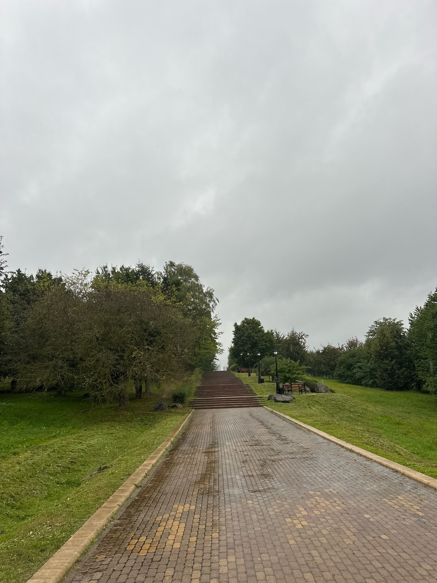 The image depicts a paved pathway leading towards a wide set of stairs in a park. The pathway is constructed from tan and brown pavers, some of which appear wet, suggesting recent rain. It is bordered by a low concrete curb and a grassy slope. To the left of the pathway, the slope is covered in lush green grass, and further up, there are trees. To the right of the pathway, the slope is also covered in grass and appears to ascend towards a line of trees. The stairs are made of a reddish-brown material and are visible at the top of the pathway, leading upwards into the park. Several lampposts and park benches are positioned along the right side of the pathway and stairs. The sky is overcast and grey. This scene is located in Minsk, Belarus, specifically at the Stairway to Heaven in Gorky Park.