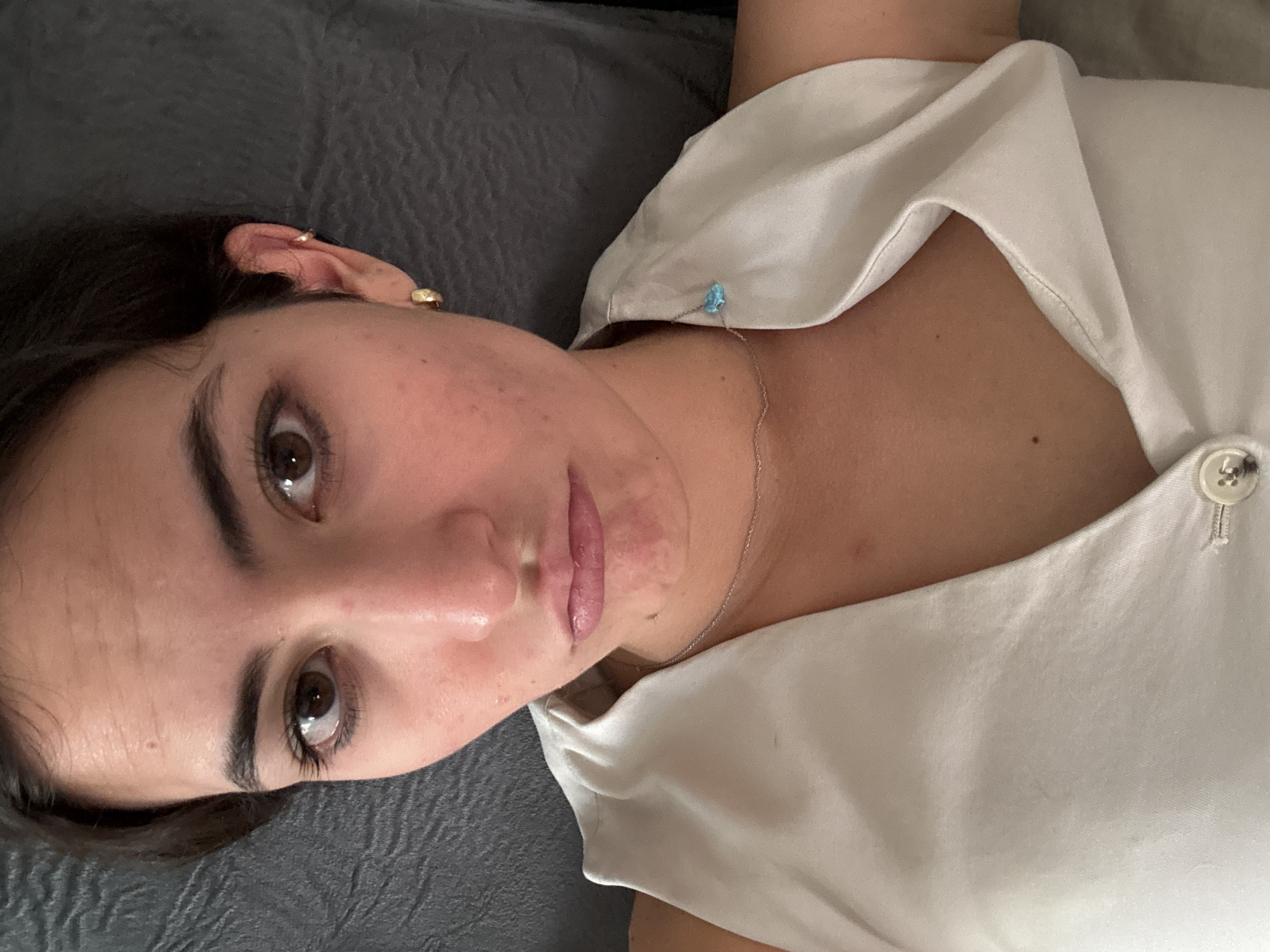 A close-up, eye-level portrait shows a young adult female with short, dark brown hair, dark brown eyes, and fair skin displaying minor facial redness and blemishes. Her lips are downturned in a pout, and she gazes directly at the camera. She wears a light-colored, likely off-white, sleeveless top or vest, partially fastened by a visible white button. A thin silver-colored chain necklace with a small, light blue, irregularly shaped charm adorns her neck. Her left ear features a small gold-colored hoop earring and a smaller gold-colored helix cuff. The background is an out-of-focus, dark grey, textured fabric. The scene is located in the city of Puebla, Mexico.
