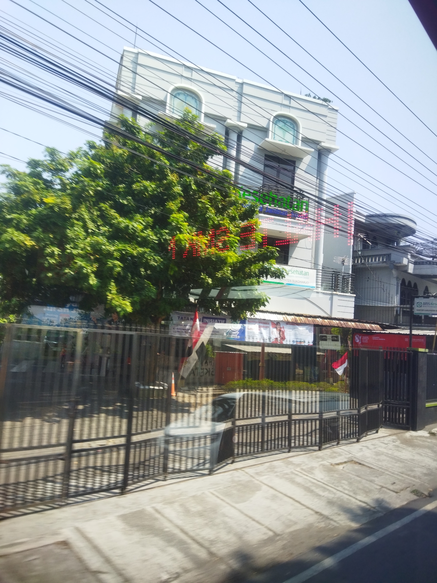 The image shows a multi-story building with a light gray facade, topped with a flat roof. The building has arched windows on the upper floors and a prominent entrance. In front of the building, there is a large, leafy green tree. Several signs are visible, some with text that appears to be in Indonesian. A black metal fence runs along the front of the property, with a sidewalk and a street in the foreground. The sky is clear and blue. Wires crisscross the upper portion of the image, suggesting the presence of electrical or telecommunication infrastructure. A small Indonesian flag is also visible, attached to a pole. The scene appears to be a street view, possibly taken from a moving vehicle. The building appears to be a commercial or administrative establishment, given the signage.