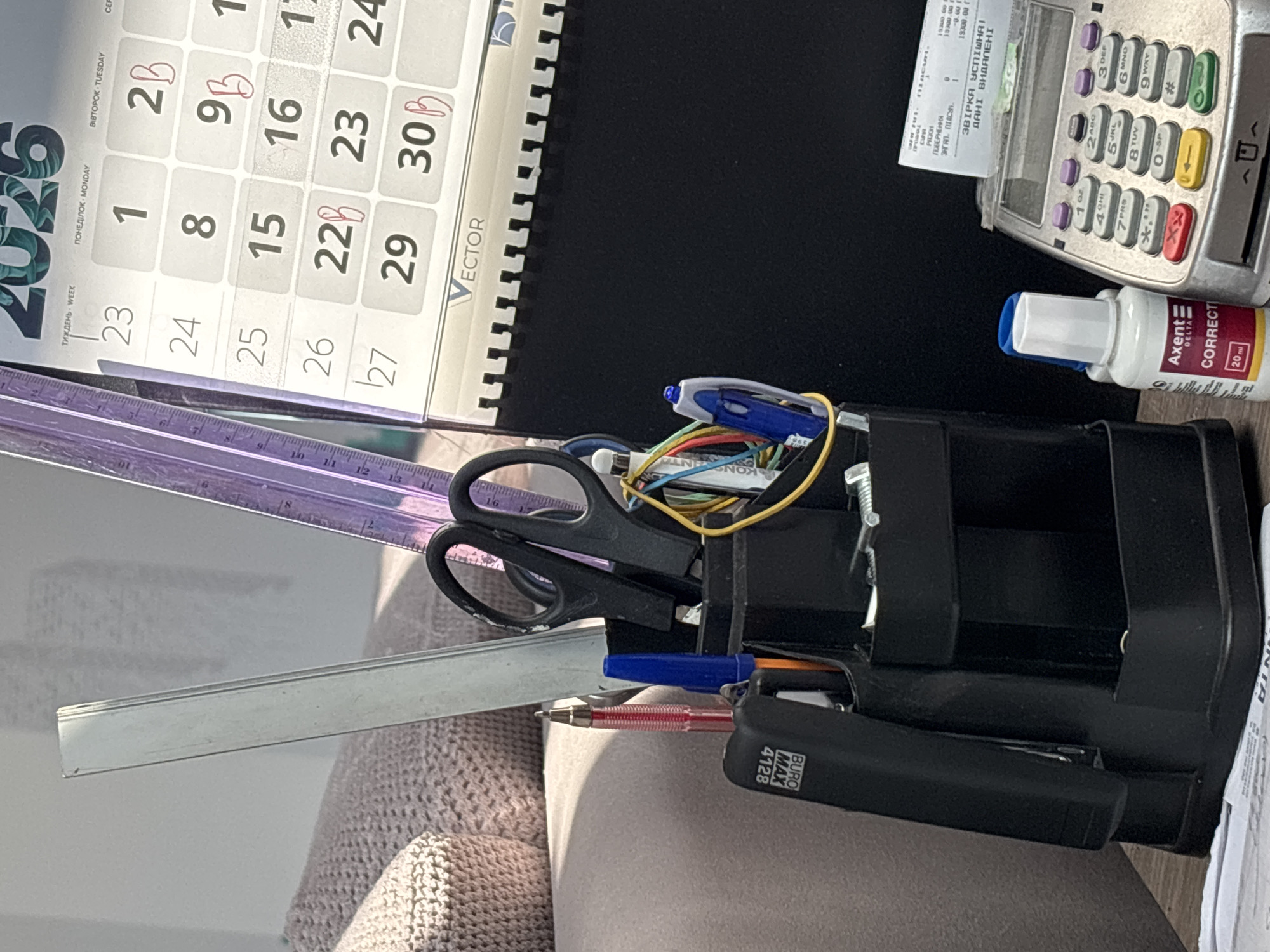 This is a close-up, overhead shot of a desk surface. The primary subjects are office supplies, including a black pen organizer filled with various pens, scissors, and a ruler. A stapler is also visible, along with a bottle of correction fluid. In the background, a receipt is visible next to a credit card machine. A desk calendar is prominent, showing the month of August 2025, with some dates circled in red. The lighting suggests it is daytime. The overall scene depicts a workspace. Text visible includes brand names like "Axent," "BURO MAX," and "VECTOR," along with a receipt in Ukrainian. The calendar shows dates and days of the week in Ukrainian and English.