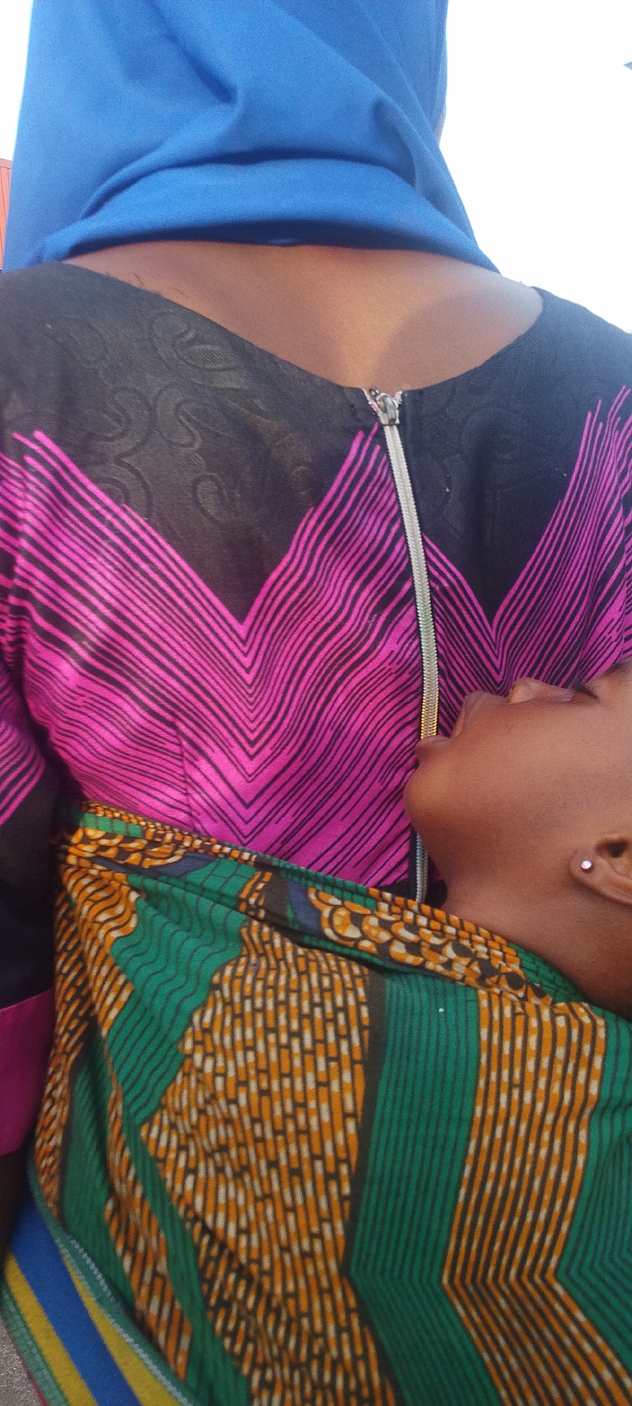 A close-up, high-angle shot captures an adult and a child in Lokoja, Nigeria. The adult, likely a woman, is partially visible from the chest and shoulders upwards. She wears a blue head covering (possibly a hijab) and a black top featuring a prominent zigzag pattern in vibrant pink stripes across the chest. A silver zipper runs down the center of her black top, extending from the collar downwards.
Nestled against the adult's chest is a child, whose face and upper body are seen in profile. The child has dark skin and appears to be resting or sleeping comfortably. A small, light-colored stud earring is visible in the child's ear. The child is wrapped or dressed in a colorful garment made of patterned fabric, predominantly green and orange with intricate geometric designs. The overall lighting is bright, suggesting daytime, though specific outdoor or indoor cues are indistinct due to the close framing. The image conveys a sense of closeness and comfort between the two individuals. There is no visible text.
