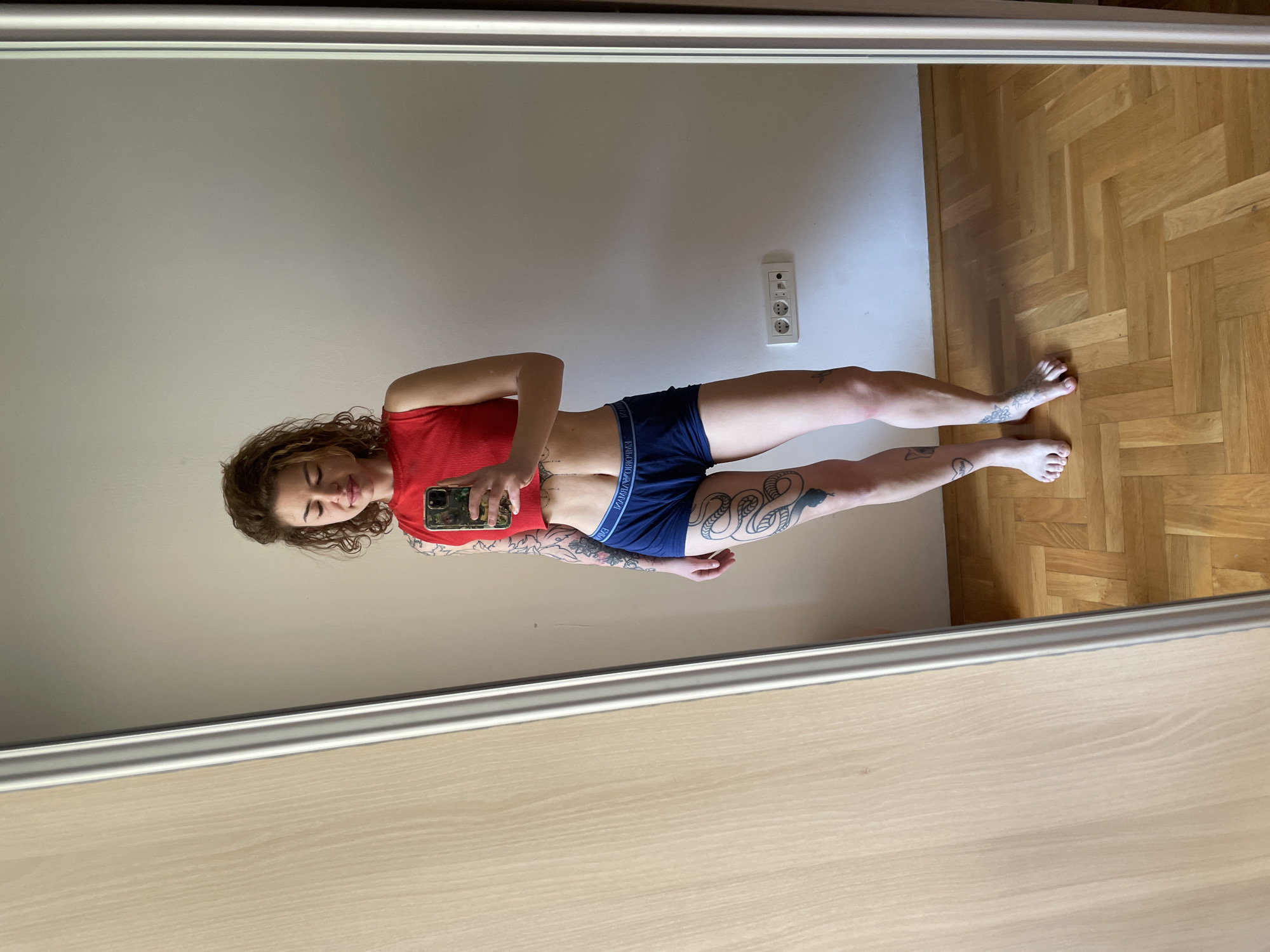 The image shows a person, presumably female, reflected in a mirror. She is wearing a red top and dark blue underwear. The subject has tattoos on her arms and legs. She is holding a mobile phone in her hand, apparently taking a selfie. The background includes a beige wall with a European-style electrical outlet and a light-colored wooden floor with a parquet pattern. Based on visual assessment of the interior and the general aesthetic, the location could be a residential setting. There is no information to pinpoint the specific city in Montenegro or whether the image was even taken there.