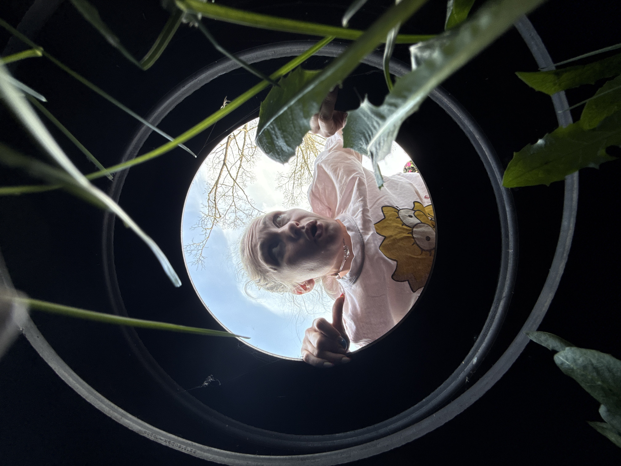 A person looking into a black plastic well or container. The person has pale skin and blonde hair and is wearing a pink shirt with a yellow cartoon character. The person appears to be giving a thumbs up. The background of the image shows a blue sky with the bare branches of a tree. The location and details of the city of Nizhnyaya Chermoda, Russia are not known.