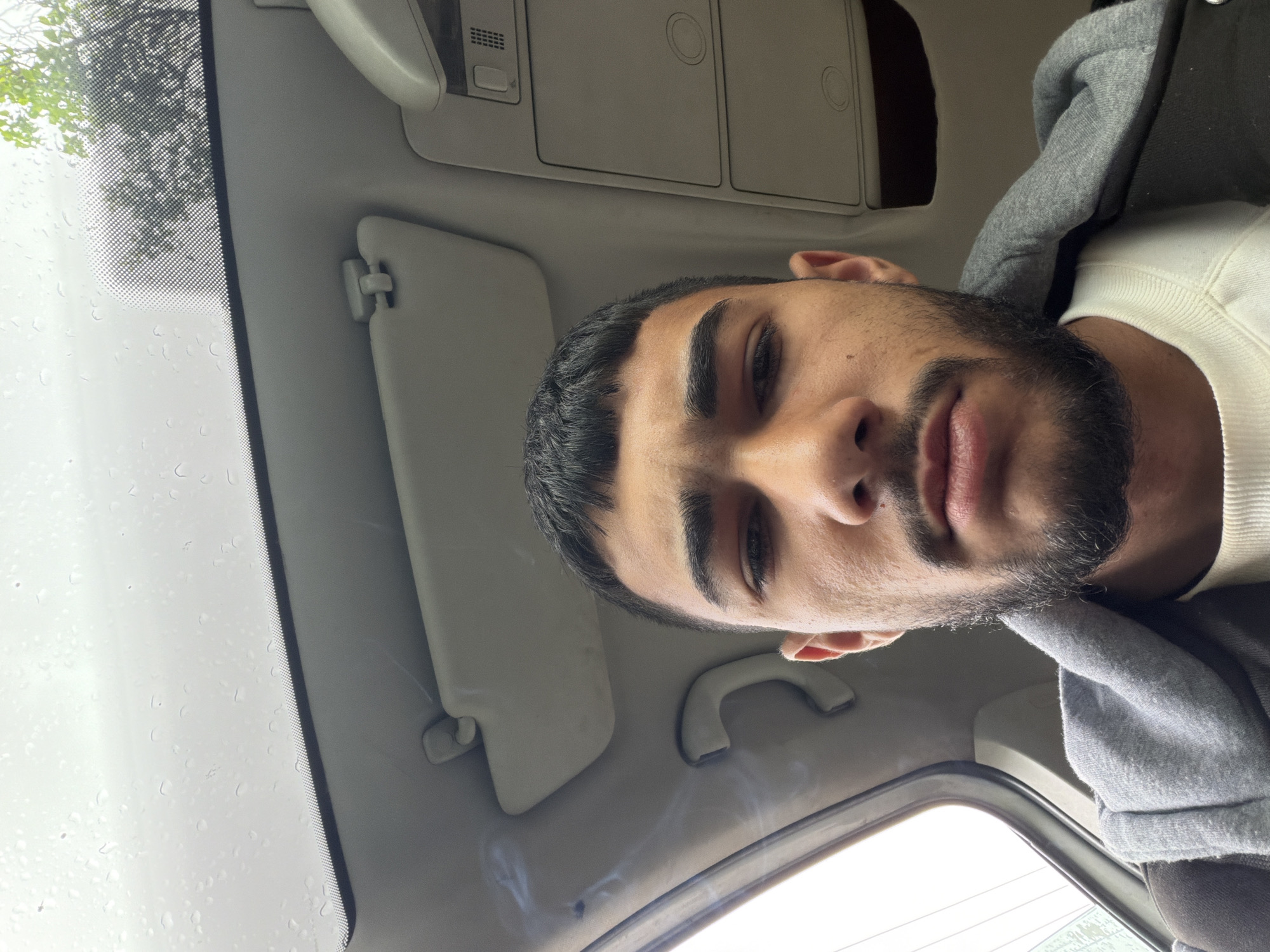 A man with dark hair and a beard is inside a vehicle, possibly a car, judging by the interior features such as a sun visor and the dashboard area visible in the frame. The vehicle's windshield shows signs of rain or water droplets. It is unclear if the man is driving or a passenger. There are no visible indicators to confirm the location as Doirentsi, Bulgaria. No further information can be derived from the image.