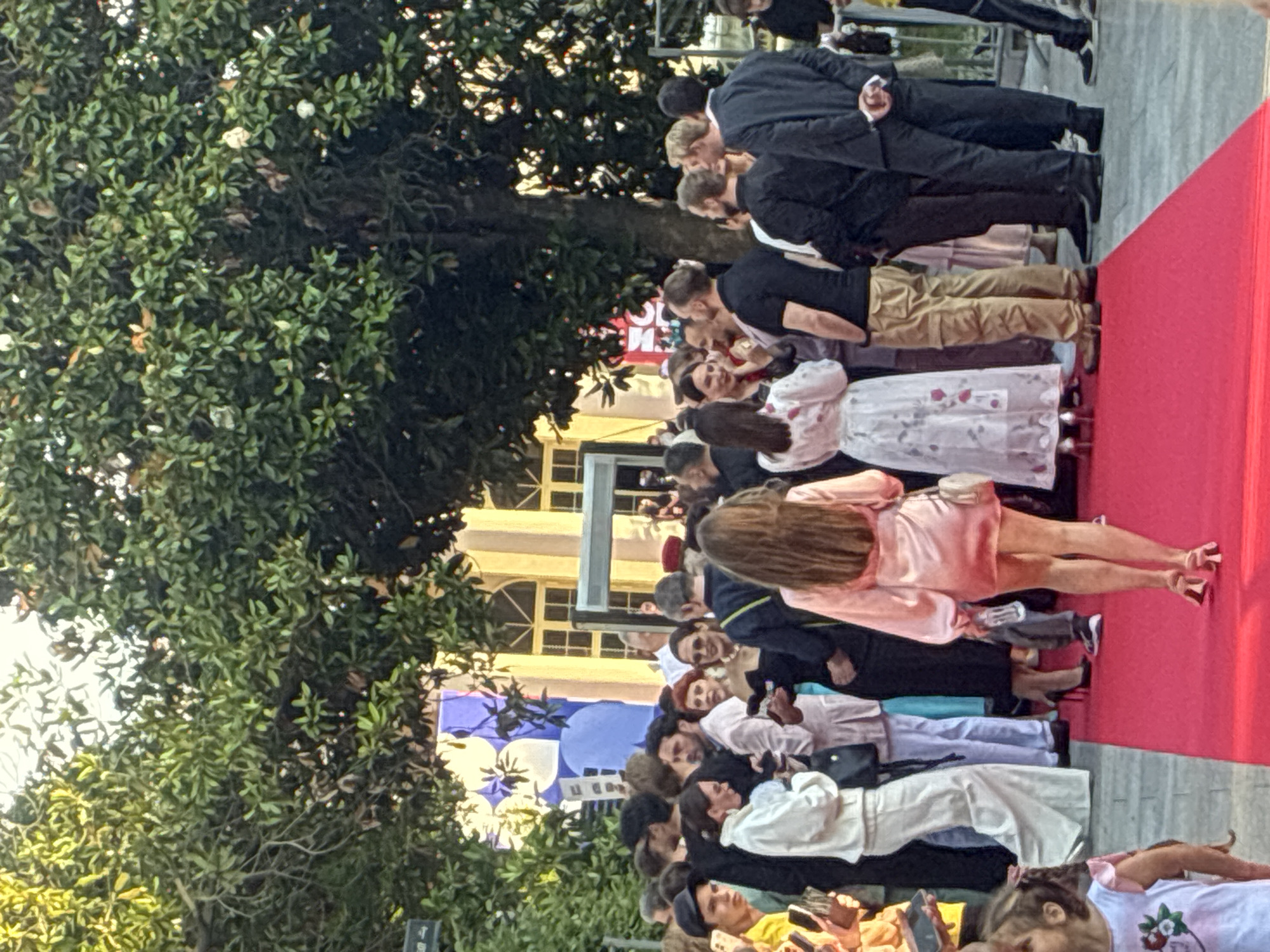 On a red carpet in Sochi, Russia, a crowd of people are gathered outdoors under the shade of large trees. Several individuals in the foreground and midground are dressed in formal attire, including suits and dresses, suggesting a special event such as a film premiere or gala. A woman in a light pink satin dress is walking away from the camera down the red carpet, holding a clear bottle in her right hand. She is wearing pink heels. In the background, a building with yellow accents and windows is visible. The atmosphere appears to be festive and public, with attendees interacting and posing for photos.