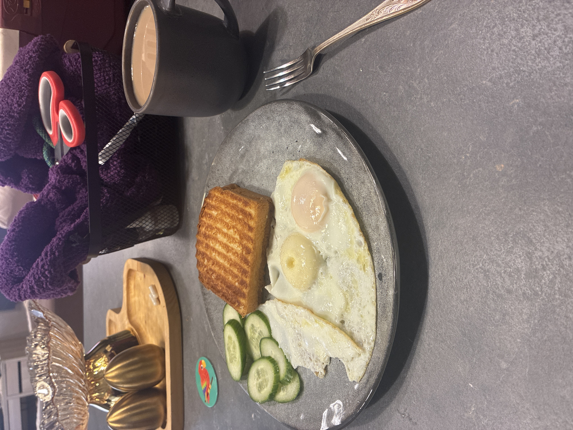An overhead shot shows a breakfast setting on a dark grey speckled countertop. In the foreground, a grey ceramic plate holds a breakfast of two fried eggs, several slices of cucumber, and a piece of toasted bread. To the right of the plate, a silver fork with an ornate handle rests on the countertop. Behind the plate, a dark grey mug filled with a light brown liquid, likely coffee or tea, is positioned. Further back, a black wire basket contains a pile of purple towels and a pair of red-handled scissors. To the left of the basket, two golden, egg-shaped decorative items sit on a light-colored wooden tray, which also holds two small white capsules. A clear glass bowl with a golden base sits behind the golden egg-shaped items. A small, colorful sticker of a parrot is placed near the plate of food. The background is slightly out of focus, showing kitchen cabinetry.