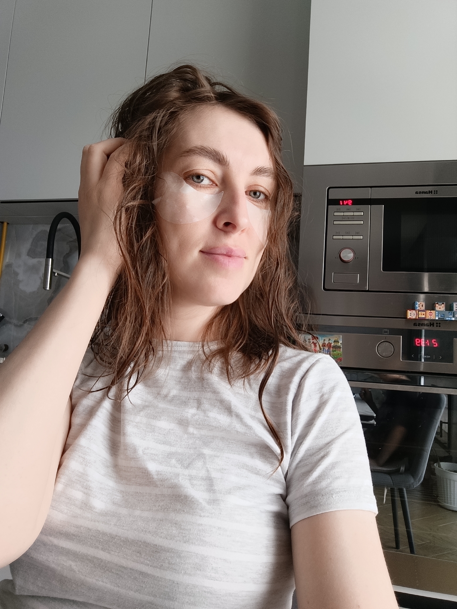 A woman is pictured indoors, presumably in the city of Chaikovsky, Russia, given the Hansa brand microwave. She is wearing under-eye patches and a striped t-shirt. Her hair appears wet and she is touching it with her left hand. Behind her is a kitchen counter, a built-in Hansa microwave and oven, and a kitchen faucet. There is a chair visible in the reflection of the oven door. The time displayed on the microwave is 1:42, and the oven display shows 8:15. Magnets are affixed to the oven.