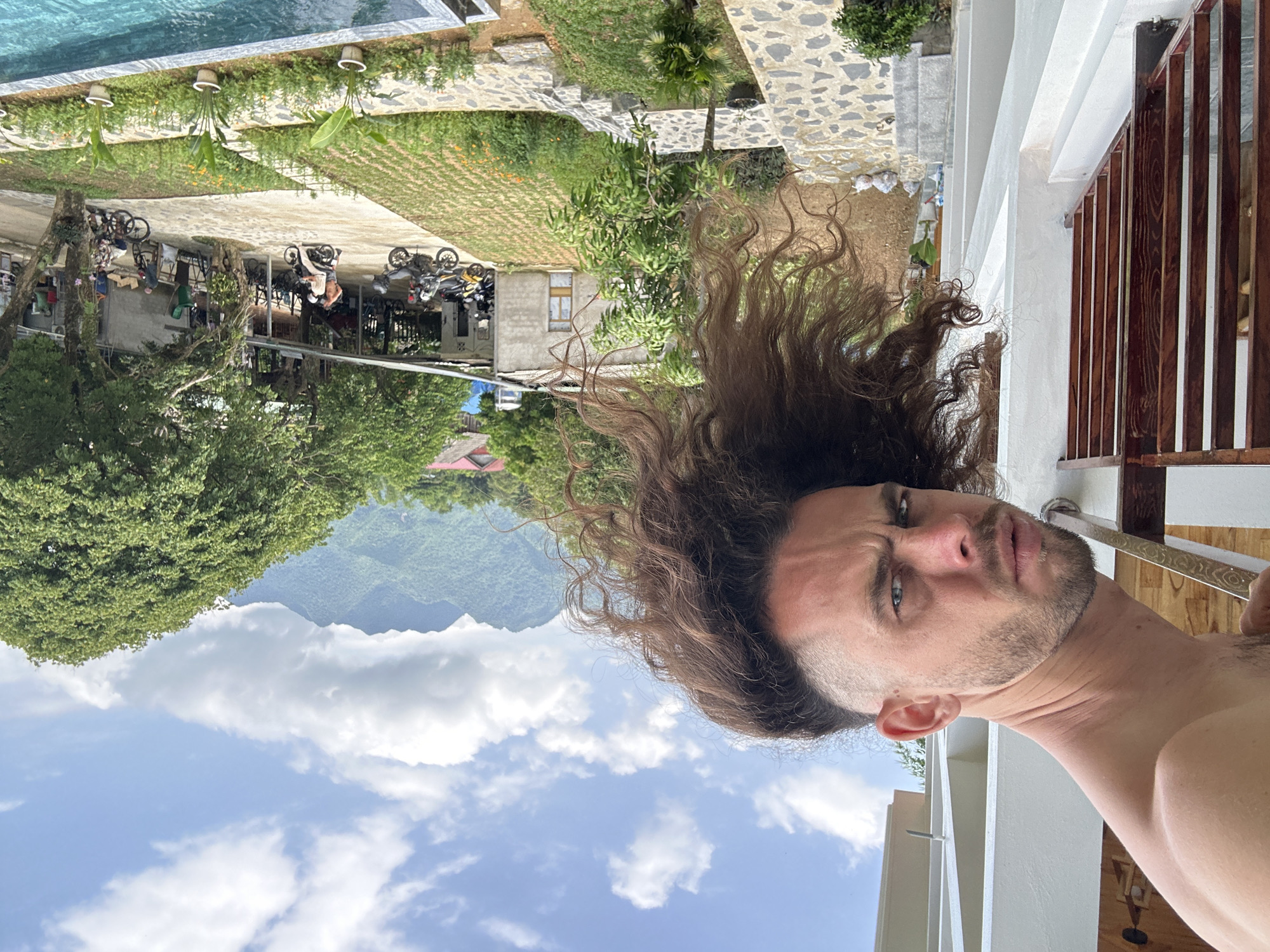 An over-the-shoulder shot captures a man with long, curly brown hair and a shaved undercut leaning over a white balcony railing. He has blue eyes and a beard, and his face is turned towards the camera with a contorted expression. The balcony is made of white plaster and has a dark brown wooden railing. In the background, a lush green landscape unfolds under a partly cloudy blue sky. Mountains rise in the distance, covered in dense foliage. Below the balcony, a courtyard features a small garden with rows of plants, stone pathways, and several motorcycles parked under a covered area. People are visible in the courtyard, some near the motorcycles, and others walking. A swimming pool with clear blue water is in the foreground, partially obscuring the view of the courtyard. The scene appears to be set in Xóm Khán, Vietnam, a rural area known for its natural beauty and mountainous terrain.
