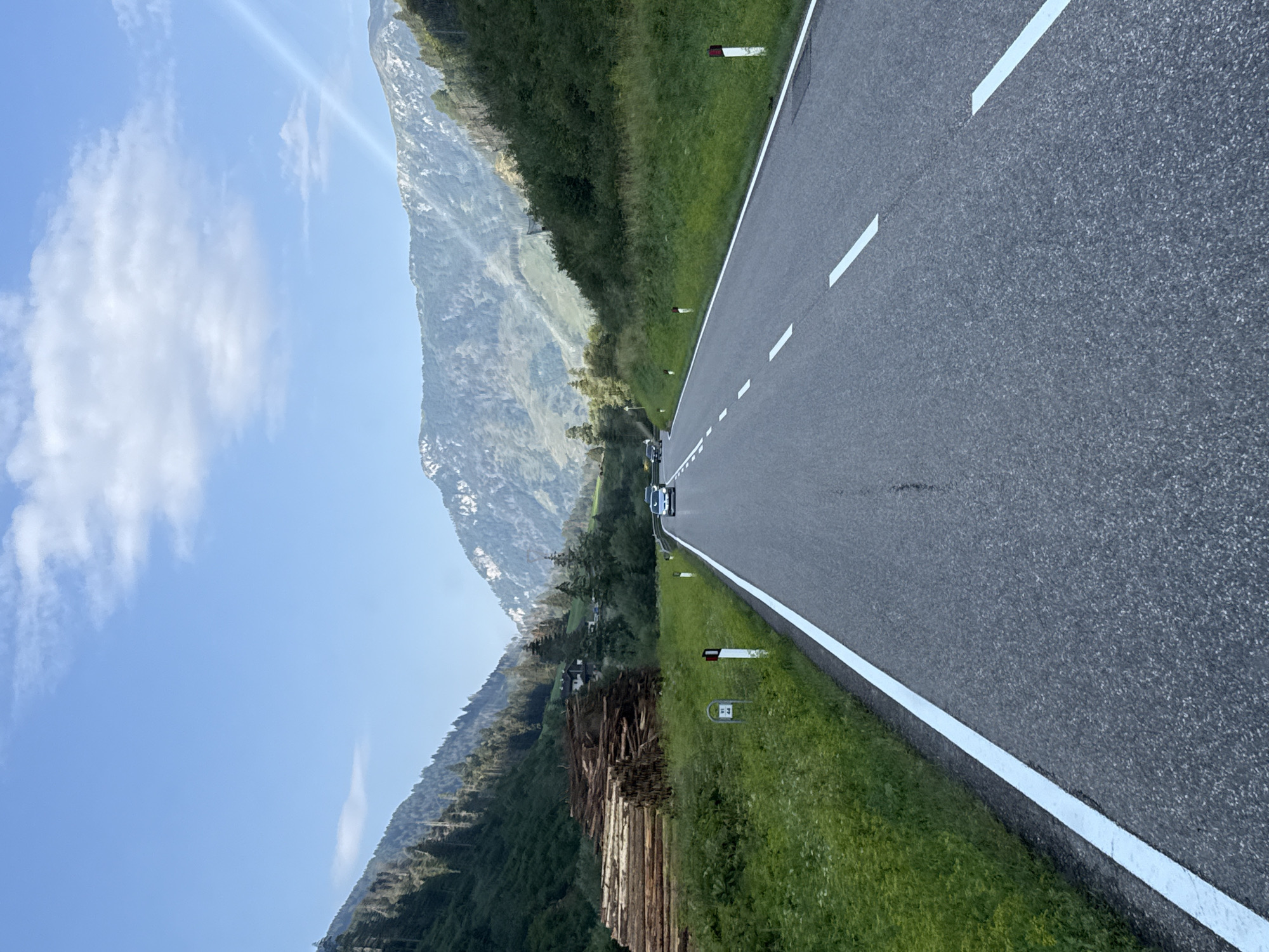 This image depicts a scenic mountain road in a rural area, likely within the municipality of Moena, Italy, known for its Dolomite mountain landscapes. The road is paved and appears to be a two-lane highway with a white solid line on the right edge and dashed white lines separating traffic lanes. The road curves gently to the right as it ascends into a valley.
On the left side of the road, a large pile of cut logs is stacked neatly, suggesting forestry activity in the area. Beyond the logs, a dense green forest covers the steep mountainside, dotted with scattered buildings, possibly homes or farm structures. The right side of the road is bordered by a grassy verge with white posts marking the roadside. Further up the mountain on the right, more dense green vegetation covers the slopes, with rocky outcrops and patches of snow visible at higher elevations.
The sky is a clear blue with a few white clouds, and sunlight beams diagonally from the upper right corner. Two cars are visible on the road, one white and one dark, driving in the same direction as the viewer. The overall environment is one of natural beauty, with towering mountains, lush greenery, and a sense of tranquility.