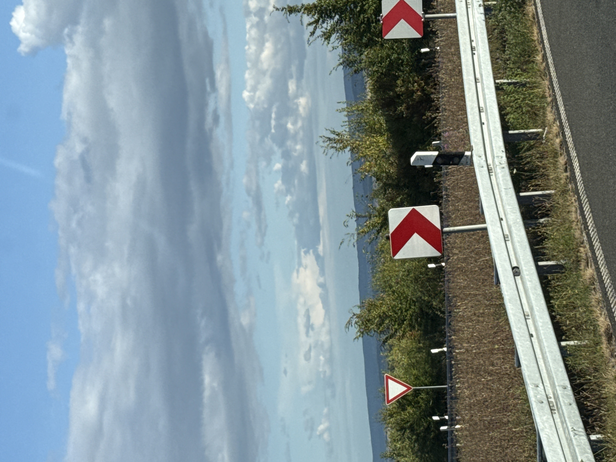 The image depicts a roadside scene in Sonnenstein, Germany, featuring a road with a metal guardrail. Several traffic signs are visible, including a red and white triangular yield sign and two white rectangular signs with red arrows indicating a sharp curve to the right. A black and white striped pole, likely a road marker, stands between the curve signs. The background consists of a line of green trees and hills under a partly cloudy blue sky. The foreground shows the asphalt road with white dashed lines.