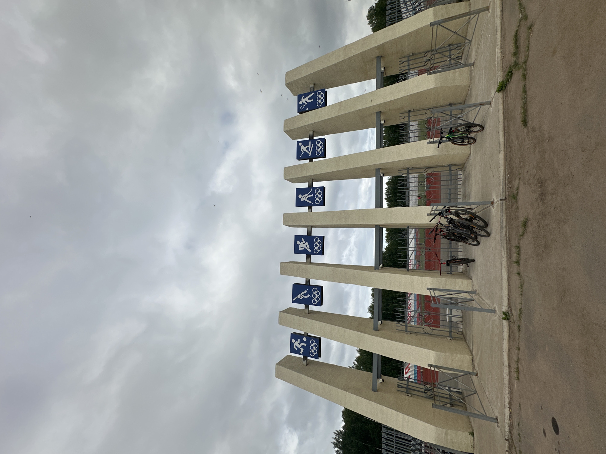 The image shows an entrance to a sports facility, likely a stadium or complex, located in the city of Chaikovsky, Russia. The entrance features a series of angled, light-colored concrete pillars, each topped with blue signs depicting Olympic sport pictograms. These pictograms include figures engaged in soccer, ice hockey, skiing, and tennis. Between the pillars are metal gates, some of which are open, revealing a glimpse of a track and seating area beyond. Several bicycles are parked in front of the entrance, indicating it's a public area. The sky is overcast with dramatic, gray clouds. The ground in front of the entrance is paved with asphalt and concrete, with some patches of sparse grass growing along the edges. The overall impression is one of a functional, outdoor sporting venue under a cloudy sky.