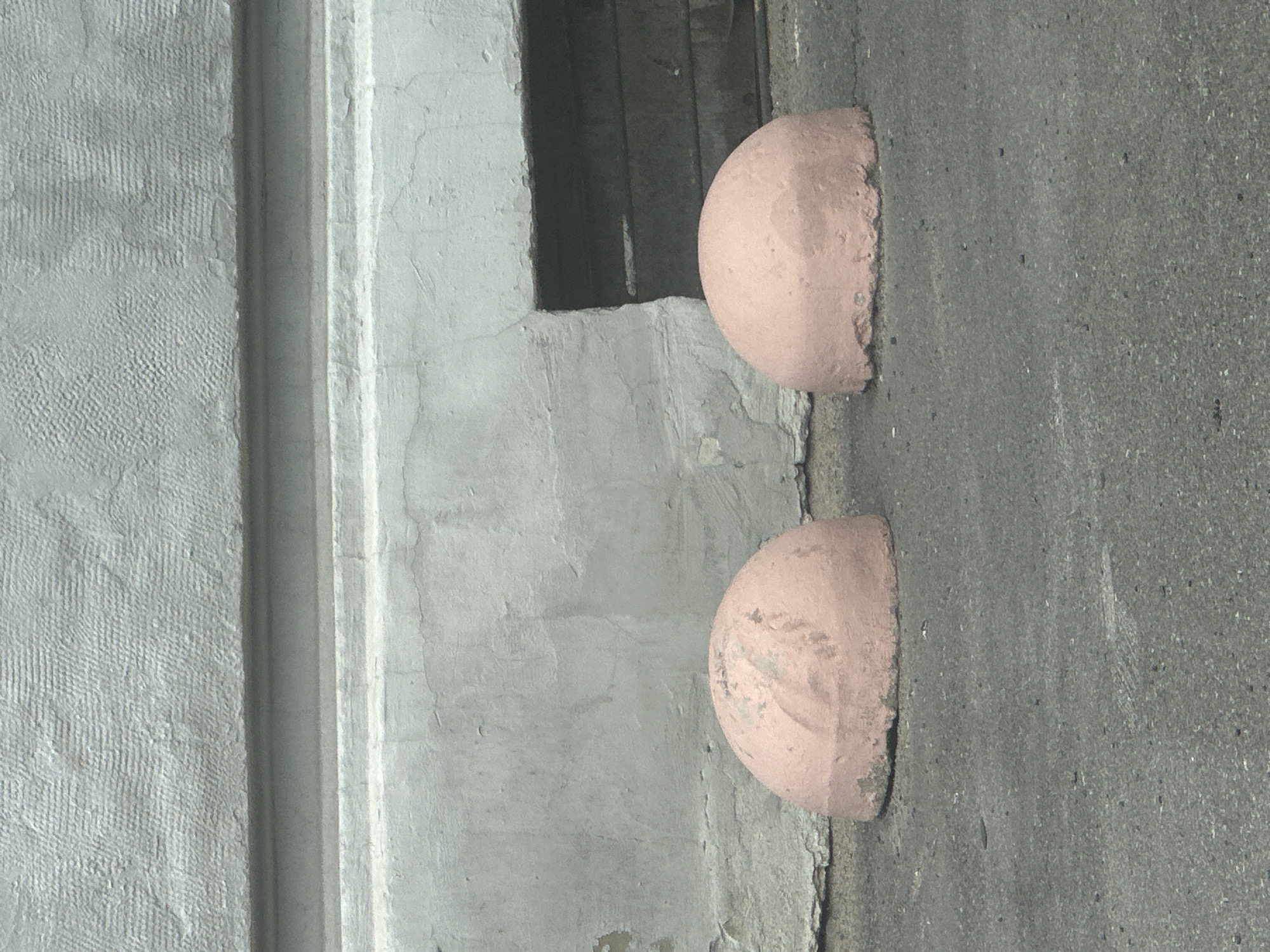 Two pink hemispherical concrete bollards are positioned on an asphalt surface in front of a light gray, textured building facade. The building has a white horizontal band separating two distinct textures. To the right of the bollards is a dark rectangular opening in the building's foundation. The overall scene appears to be an outdoor urban environment.