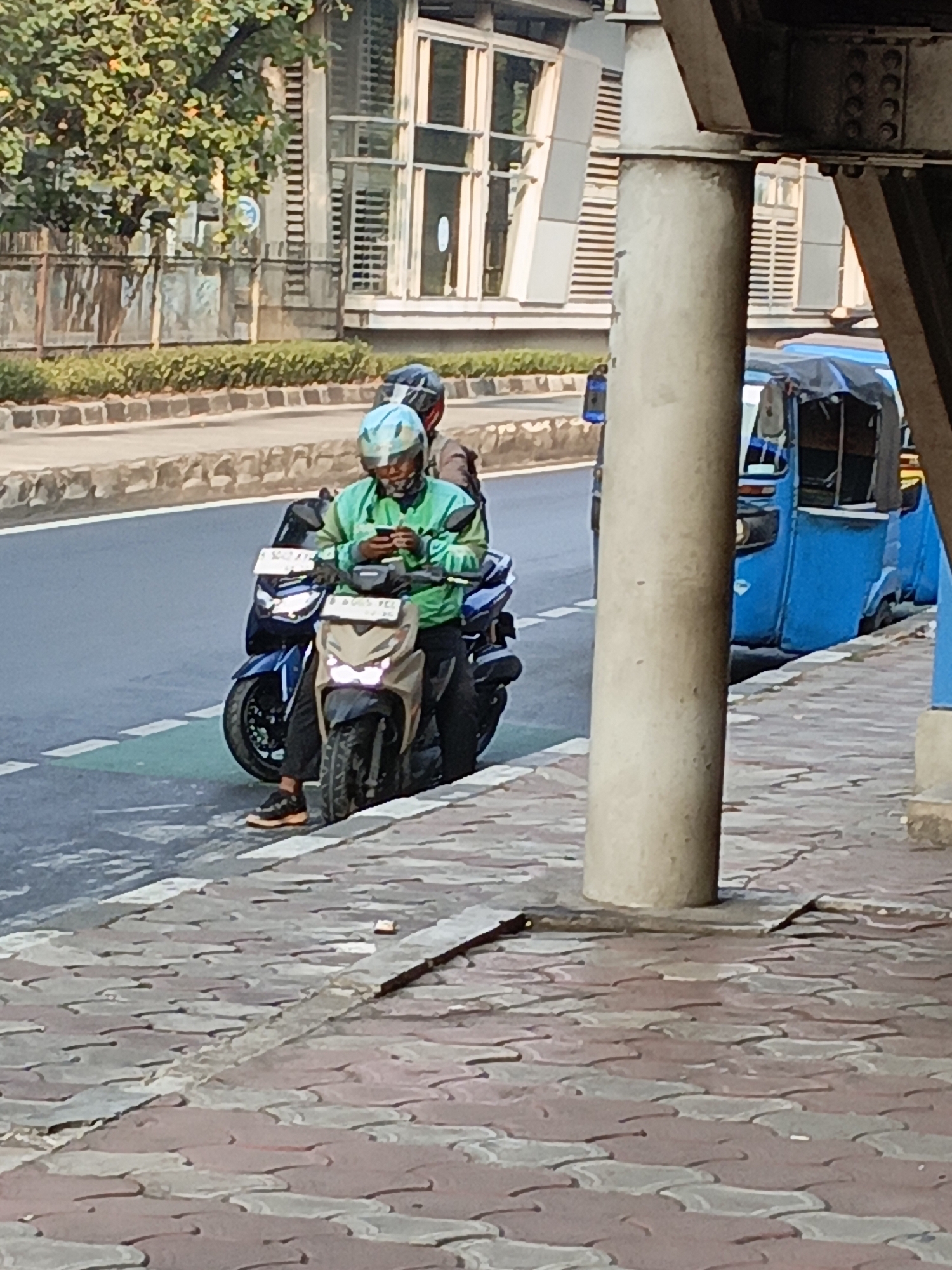 In East Jakarta, Indonesia, a person wearing a light blue and green helmet and a green jacket is seated on a light brown scooter. They are looking down at their phone, which they are holding in their hands. Behind them, another person on a blue scooter is partially visible. The scooters are parked on the side of a road, adjacent to a pedestrian walkway made of patterned paving stones. In the background, a blue auto-rickshaw is parked further down the road, and a building with large windows and blinds is visible. The scene is illuminated by daylight, casting shadows from a large concrete pillar in the foreground.