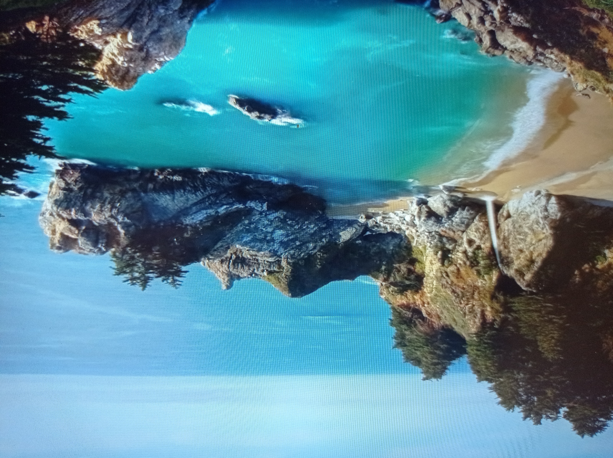 The image depicts a scenic coastal landscape with a waterfall cascading onto a sandy beach, which then meets turquoise-colored water. Rocky cliffs covered in lush green vegetation rise from the water, with evergreen trees dotting their tops. The sea stretches out to the horizon, meeting a clear blue sky. There are no people or man-made objects visible in the image, aside from what appear to be two small, distant boats. The scene is tranquil and natural, evoking a sense of secluded beauty. The location is not in Mukachevo, Ukraine, but rather appears to be a natural coastal area.