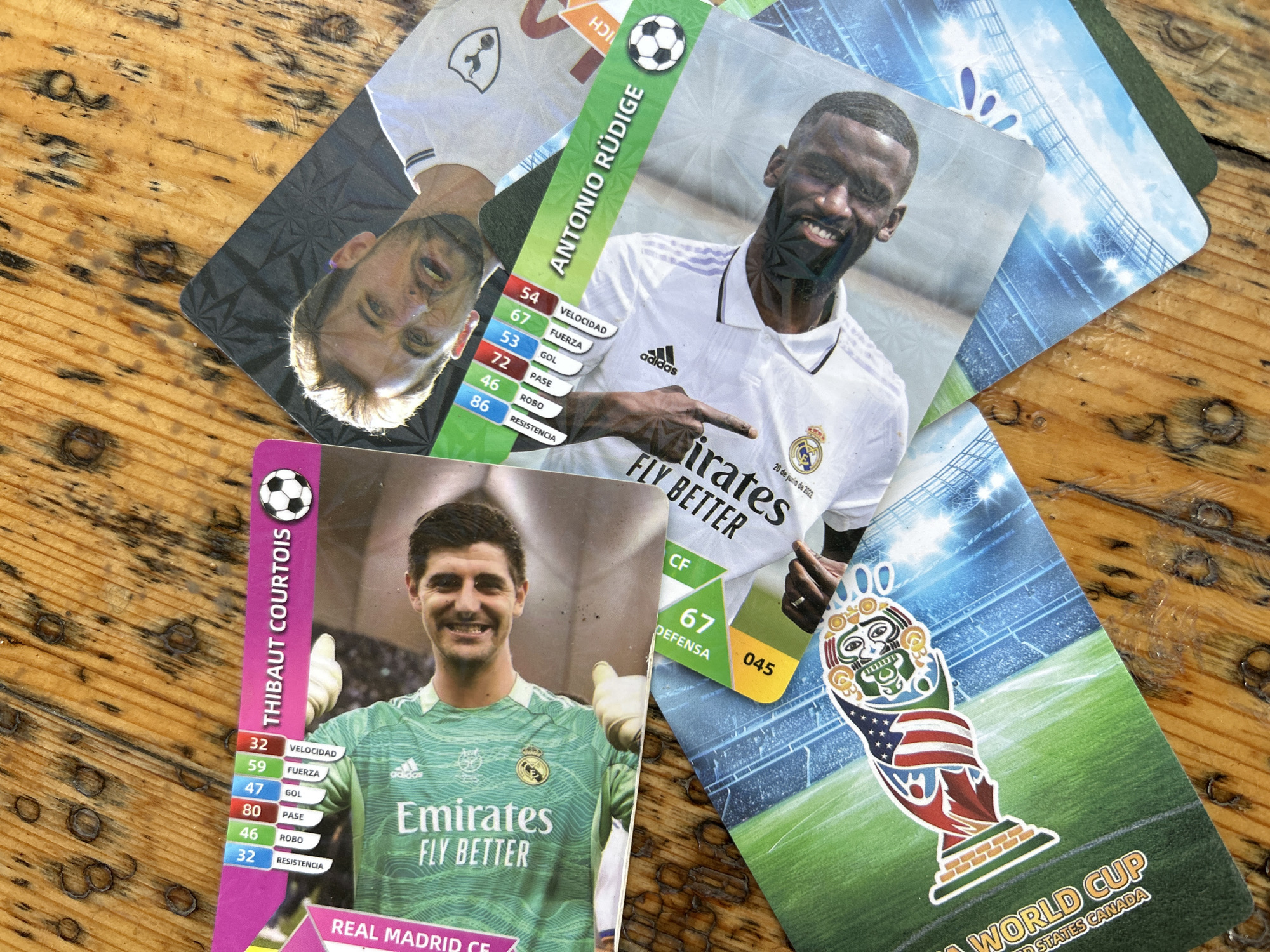 A collection of football trading cards is casually laid out on a light brown wooden surface, featuring visible grain and dark stains. The cards depict various players and football themes, presented at different angles and overlapping.
Two prominent cards feature smiling footballers. In the center, Antonio Rüdiger, wearing a white Real Madrid jersey with the adidas logo and "Emirates FLY BETTER" sponsorship, is shown. His card displays stats like "VELOCIDAD" (54), "FUERZA" (67), "GOL" (53), "PASE" (72), "ROBO" (46), and "RESISTENCIA" (86), along with "DEFENSA" (67) and the number "045". Text on his card also includes "ANTONIO RÜDIGE" and "20 de junio de 202".
Below Rüdiger's card, Thibaut Courtois is featured, also smiling and wearing a green Real Madrid goalkeeper kit with "Emirates FLY BETTER" and the adidas logo. His card lists "THIBAUT COURTOIS" and stats: "VELOCIDAD" (32), "FUERZA" (59), "GOL" (47), "PASE" (80), "ROBO" (46), and "RESISTENCIA" (32), plus "REAL MADRID CF".
Partially visible in the top left is another card showing an unidentified player's face, wearing a white jersey, with the text "HDI". Another card in the bottom right corner features a stylized World Cup trophy incorporating elements of the United States and Canadian flags, against a stadium backdrop. Text on this card reads "WORLD CUP" and "UNITED STATES CANADA", suggesting the 2026 FIFA World Cup. The cards generally have a vibrant, colorful design, with football imagery and player statistics highlighted.