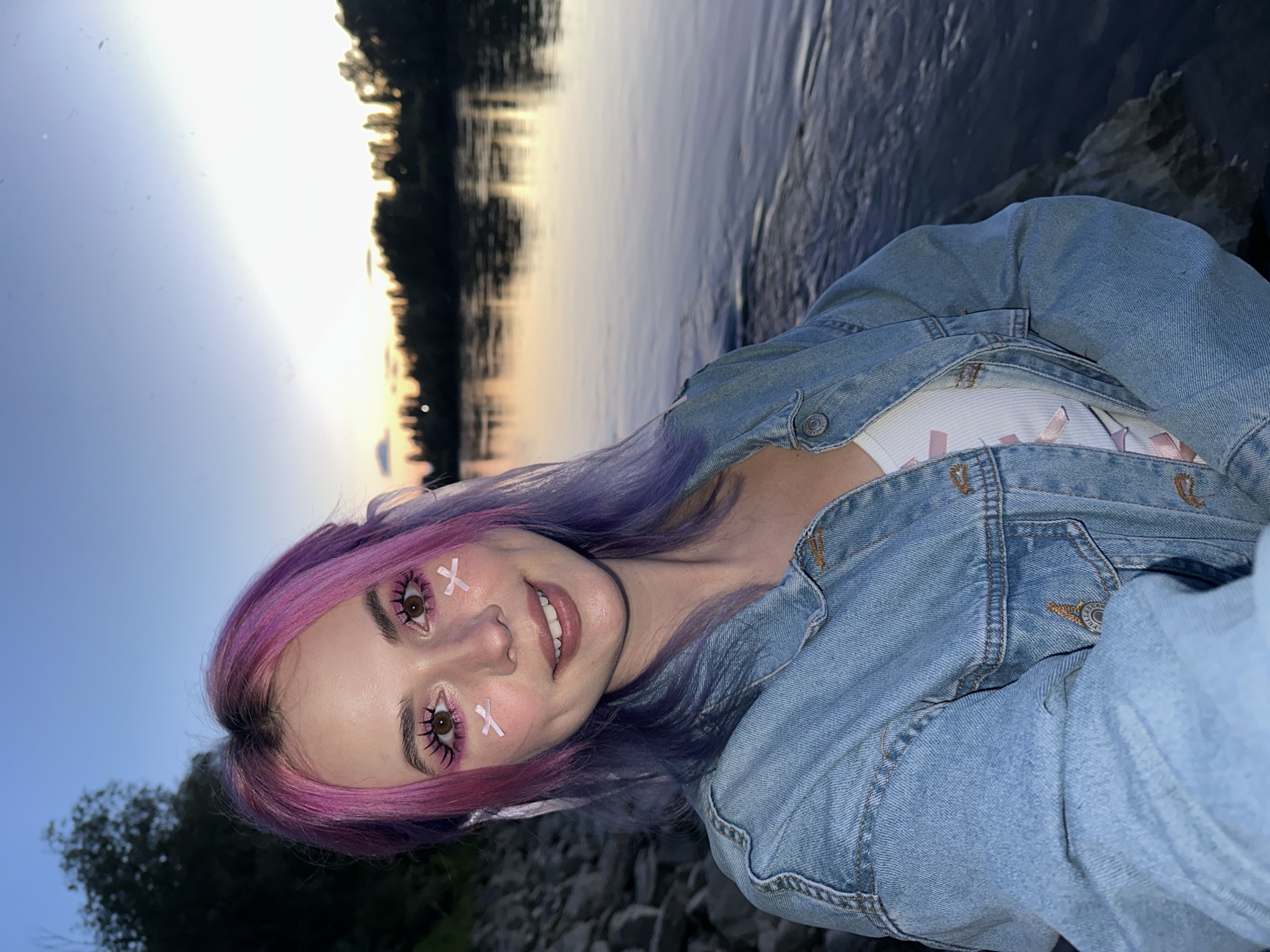 A young woman with dyed pink and purple hair poses for a photo in an outdoor location. She is wearing a light blue denim jacket over a white top with pink accents. Her makeup includes pink eyeshadow and small pink "x" marks near her eyes. The background features a body of water, likely a lake or river, with trees lining the opposite shore. The lighting suggests it is either sunrise or sunset. The scene does not depict any specific identifiable landmarks within Zagreb, Croatia, nor is there enough data to suggest the image was taken there. The woman is smiling slightly. The ground appears to be rocky or pebbled. The image is rotated slightly clockwise.