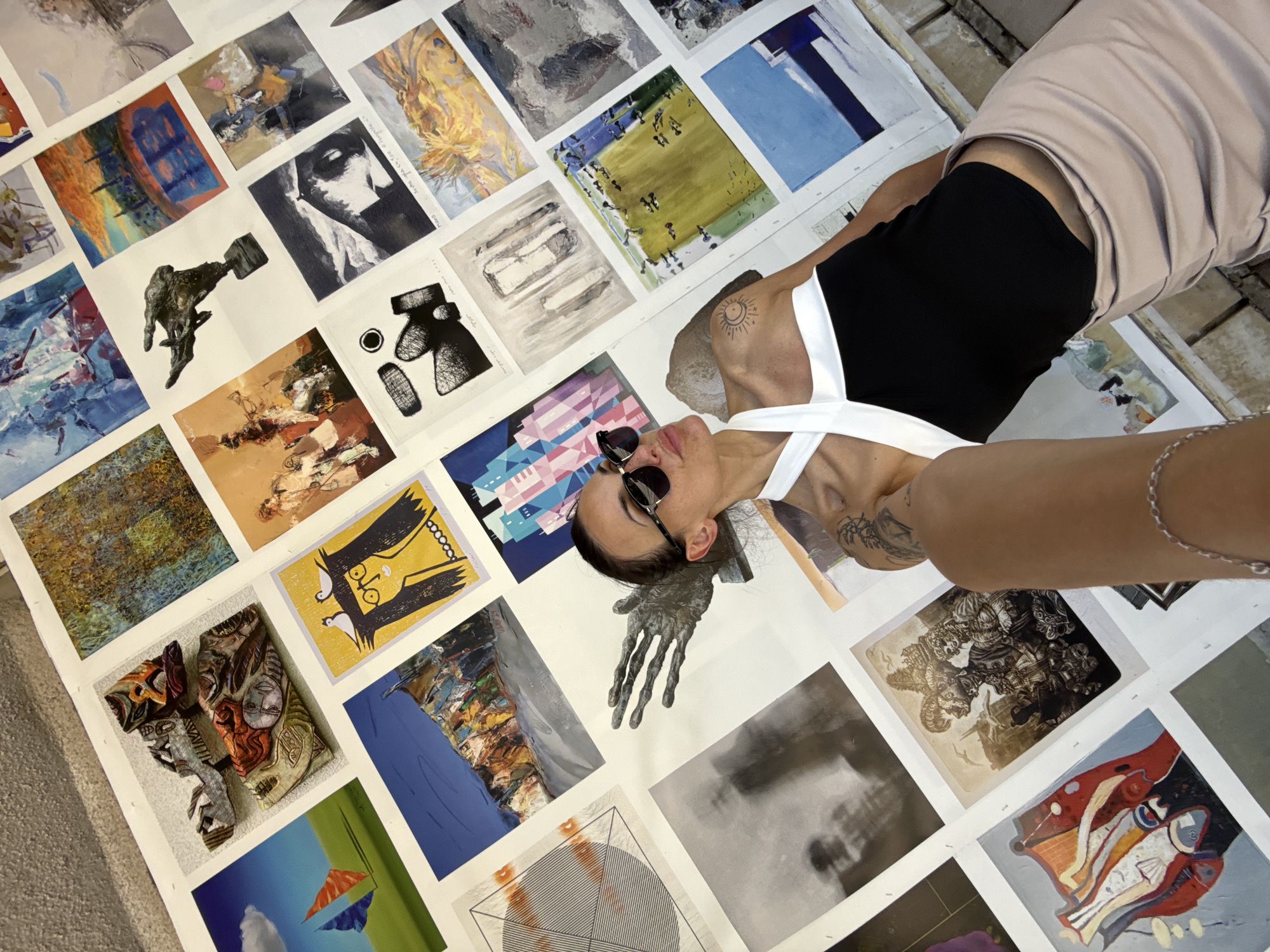 A woman lies on a large arrangement of art prints, presumably in Pleven, Bulgaria. She wears sunglasses, a black top with white straps, and a light brown skirt. Her hair is pulled back. She has tattoos on her shoulder and arm, and a silver chain bracelet on her wrist. The art prints vary in style and subject matter, including abstract designs, portraits, and figures. The background appears to be a stone or concrete floor, possibly outdoors. Her left hand is also replicated in an art piece next to her head.