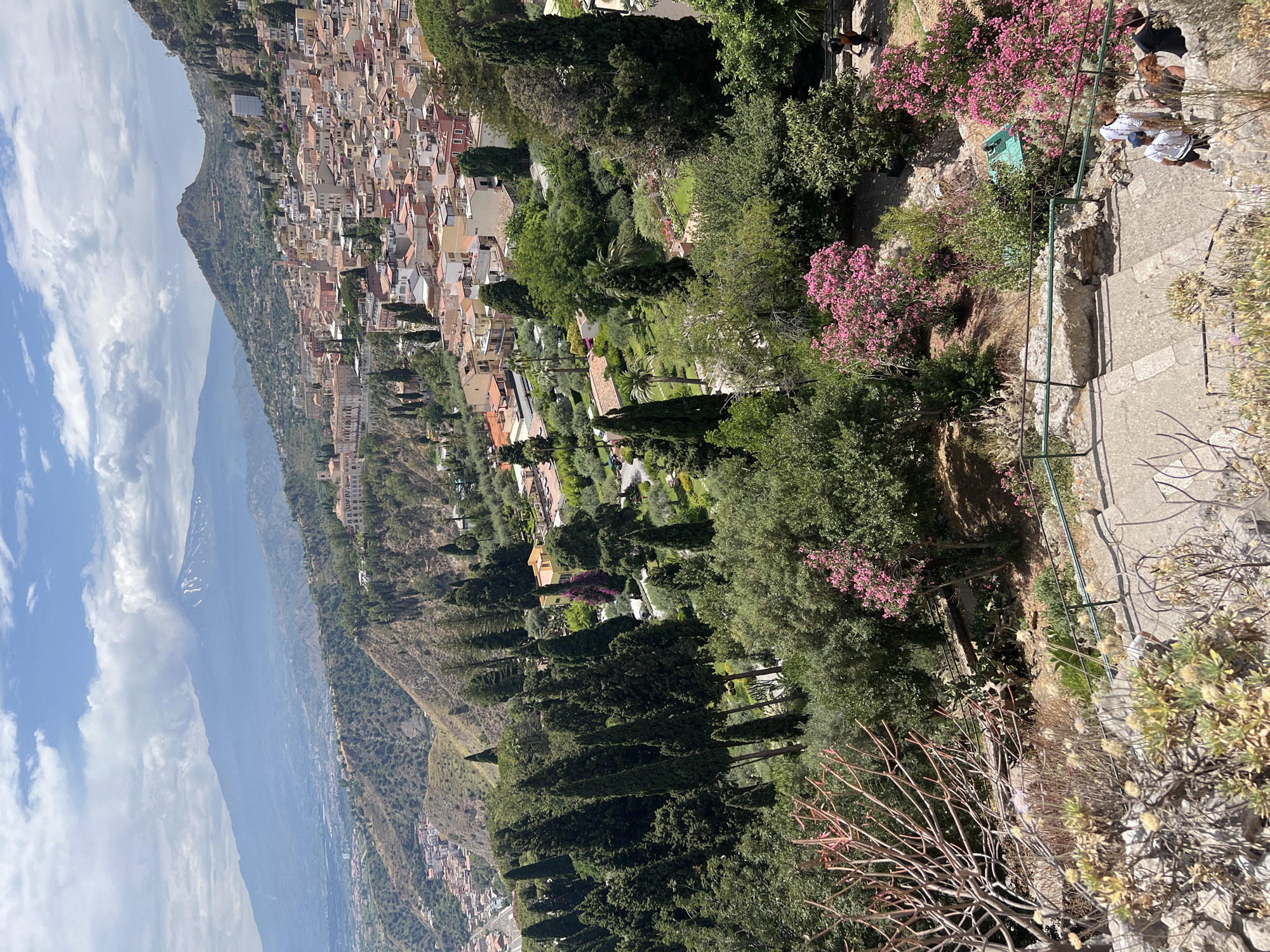 This image, captured in Taormina, Italy, showcases a panoramic view from a hillside. The foreground features a stone staircase with a green metal railing, flanked by dry, rocky ground and sparse vegetation, including succulents and bare branches. Several individuals are seen descending the stairs. Beyond the stairs, a lush garden unfolds, characterized by manicured lawns, trees, and vibrant pink flowering bushes. In the middle ground, a densely populated town with terracotta-tiled roofs climbs the hillside, interspersed with cypress trees. Further in the distance, rolling hills give way to a hazy, snow-capped mountain, likely Mount Etna, under a partly cloudy sky.