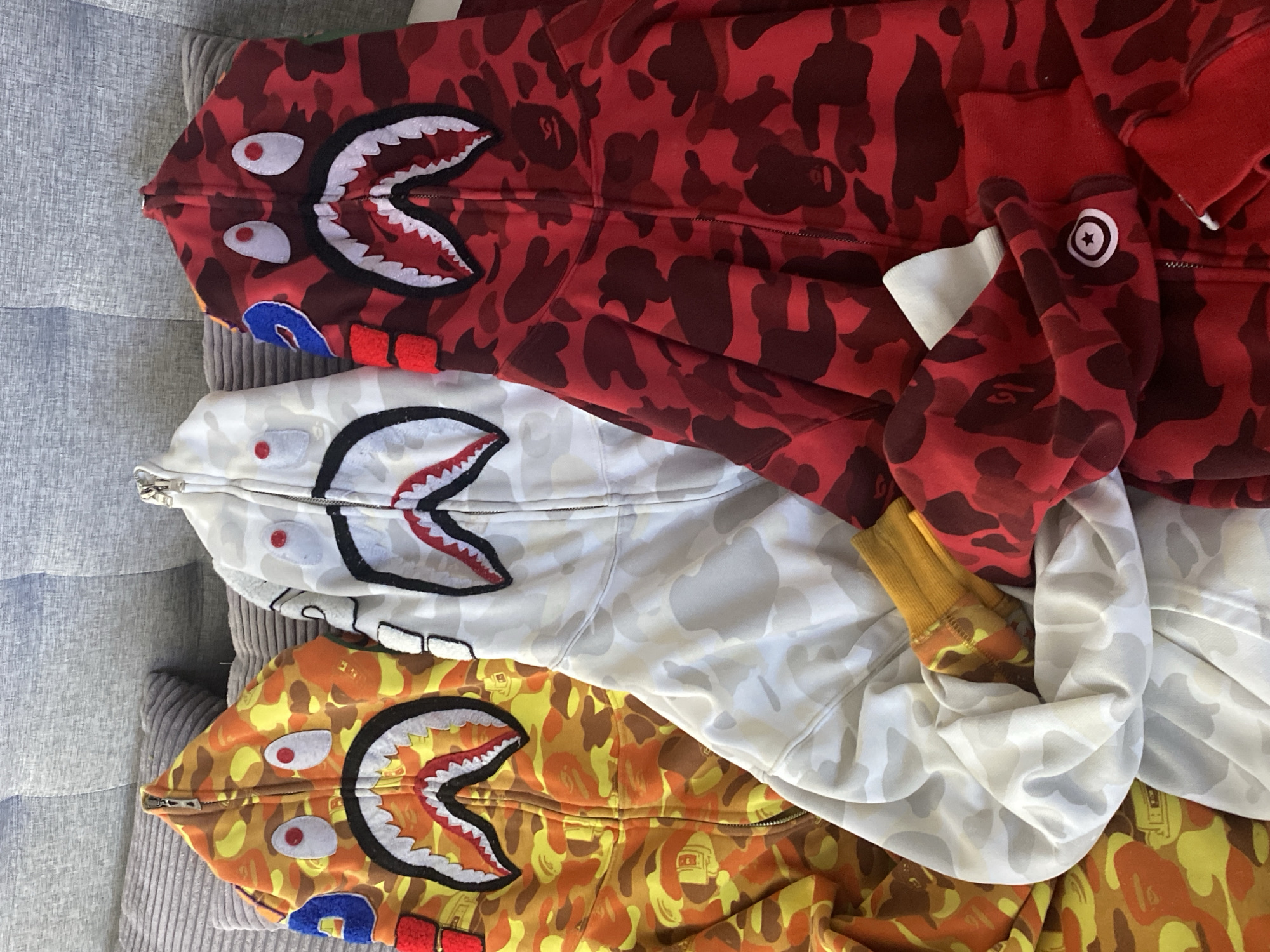 The image displays three A Bathing Ape (BAPE) full-zip "shark" hoodies, neatly laid out and partially overlapping on a grey, ribbed upholstered surface, likely a sofa. The hoodies are arranged vertically, with their distinctive hoods featuring the "shark face" design prominently displayed at the top of the frame.
The top hoodie features a red camouflage pattern, incorporating the BAPE ape head logo. Its hood is fully zipped, revealing white eyes with red pupils, a sharp-toothed mouth, and blue and red fabric details resembling gills on the sides. A circular white logo with a star design is visible on the cuff of this red hoodie, along with a white fabric tag.
Below the red hoodie is a white and grey camouflage version. This hoodie also displays the iconic shark face design on its fully zipped hood, with white eyes and a black-outlined, toothy mouth with red accents.
The bottom hoodie exhibits a vibrant yellow and orange camouflage pattern, similarly integrating the ape head logo. Its hood, also fully zipped, features the shark face design with white eyes, red pupils, and a mouth full of white teeth outlined in black. Blue and red fabric details are visible on the sides of its hood.
The image captures a static display of these streetwear garments, suggesting they are being showcased or photographed. No people are visible, and no specific activities are taking place. The setting appears to be indoors, under ambient lighting. While the location context provided is Sochi, Russia, there are no visual cues within the image itself to confirm this geographical detail. The visible text includes the BAPE ape head within the camouflage patterns and the star logo on the red hoodie's cuff.