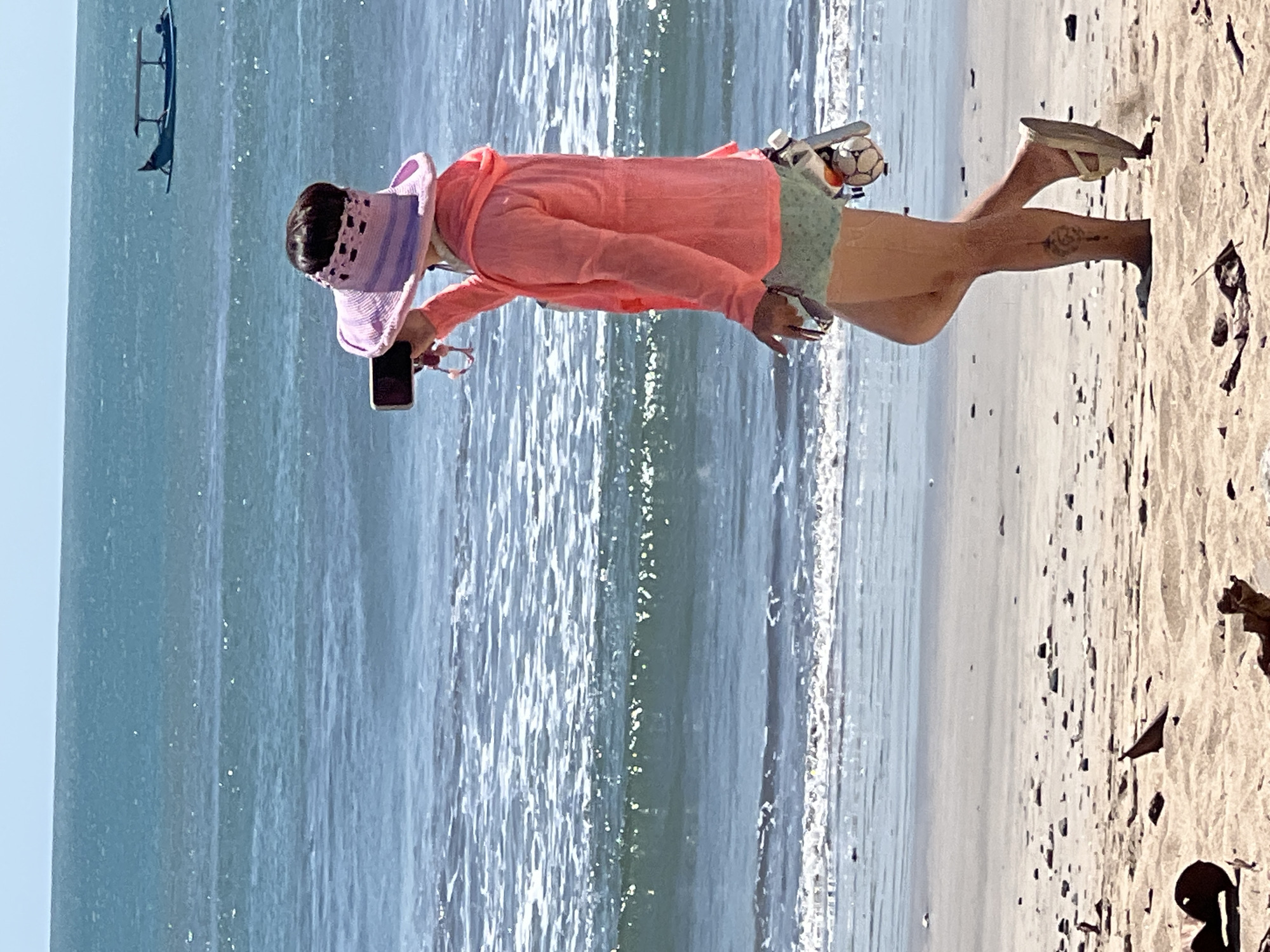 A woman is walking on a sandy beach in Kuta, Indonesia, with the ocean behind her. She is wearing a pink sun hat with purple stripes, a coral-colored hooded jacket, and light green shorts. In her right hand, she is holding a smartphone to take a picture. A small, woven bag is attached to her shorts, and it contains a small, soccer ball-like object. Her left leg has a tattoo of a symbol. The ocean is blue with white waves crashing, and a small boat with a canopy is visible in the distance. The sand on the beach is light brown and littered with small debris. The sky is a clear, pale blue.