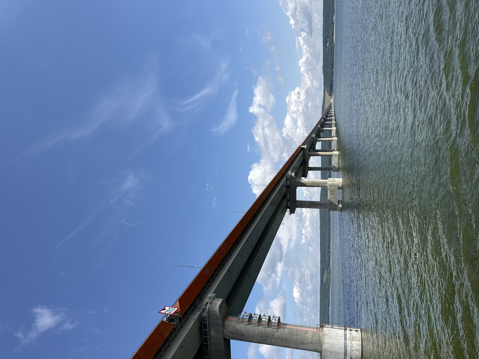 A wide shot captures a long, elevated bridge spanning a body of water under a bright blue sky dotted with fluffy white clouds. The bridge, with its orange-red top deck and concrete pillars, extends into the distance, flanked by a green, tree-covered shoreline. The water below is a murky green with gentle ripples. A sign with the number "28.6" is visible on the side of the bridge. The scene appears to be in Shelanga, Russia.