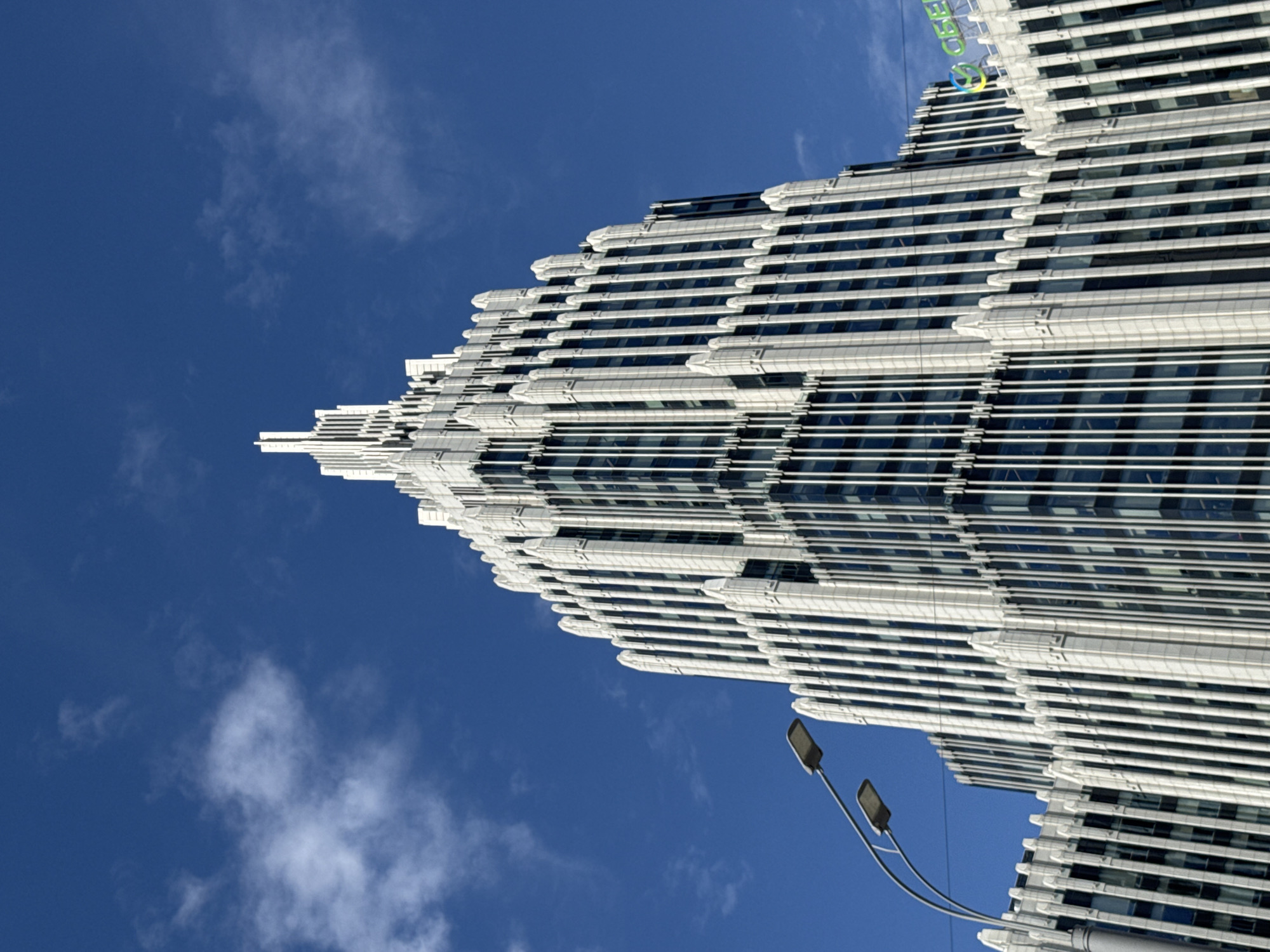 The image is a low-angle, full-frame shot of the iconic "Seven Sisters" style skyscraper, specifically the Ministry of Defense building on Frunzenskaya Embankment in Moscow, Russia. The building features a distinctive art deco design with a central spire and tiered setbacks, adorned with vertical white fins and dark glass windows. A double street lamp post with curved arms is visible in the lower left corner, extending towards the building. The sky is a vibrant blue, punctuated by scattered white clouds. The top of another building, with a green logo that reads "SBER," is partially visible in the upper right portion of the frame. The overall scene depicts a clear, sunny day with the monumental architecture of Moscow dominating the composition.