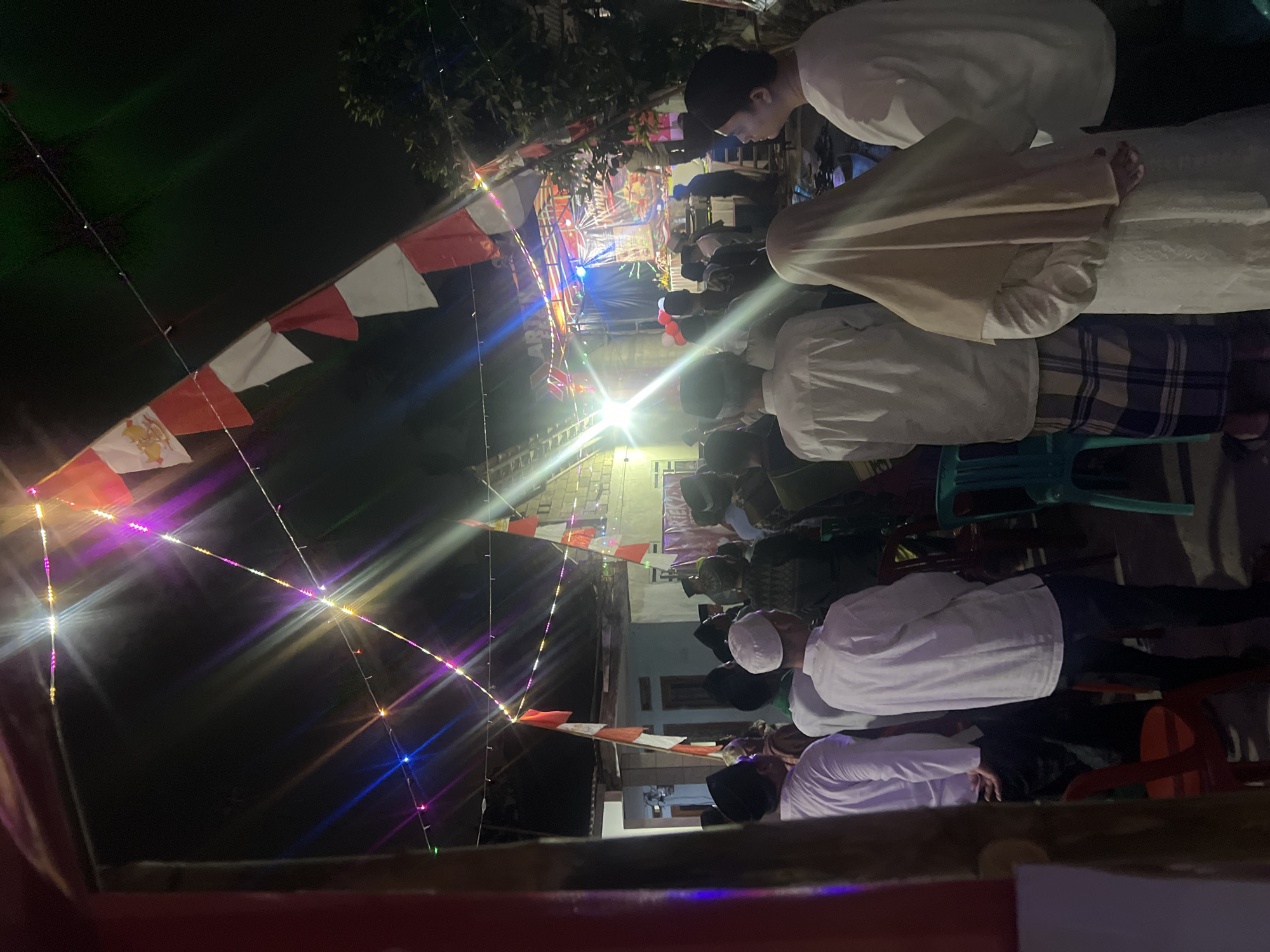 The image depicts an outdoor nighttime gathering in Klapanunggal, Indonesia. A crowd of people, many wearing traditional Islamic attire such as peci hats and sarongs, are facing a stage area illuminated by colorful lights. The stage appears to be set up for a performance or event, with a banner visible on the backdrop. Red and white Indonesian flags are strung across the scene, along with decorative string lights. The environment suggests a community event, possibly a festival or celebration, taking place in a residential or open area.