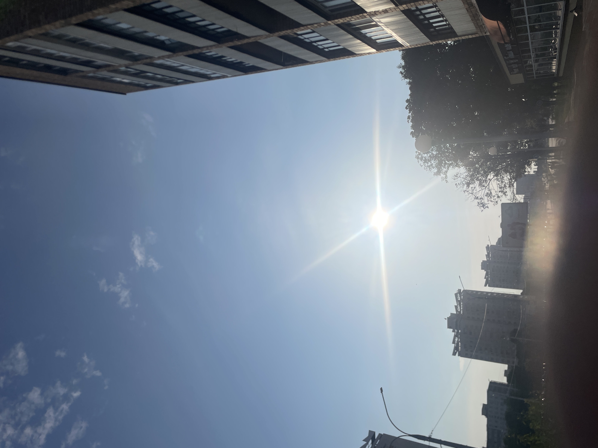 The image depicts a bright, sunny day in a city, likely in an urban or suburban setting. The sky is a clear, light blue with a few wispy white clouds scattered across it. The sun is prominently featured in the center of the frame, emitting strong lens flares that create a starburst effect.
On the right side of the image, a tall, multi-story building dominates the foreground. It appears to be made of brick and has numerous windows. Below this building, there is a storefront with a patterned awning and large glass windows.
To the left and in the background, several other apartment buildings are visible, varying in height and architectural style. Some of these buildings are silhouetted against the bright sky. Streetlights are also present, with one clearly visible in the mid-ground. Lush green trees are situated between the buildings, adding a touch of nature to the urban landscape. The lower portion of the image is blurred, suggesting it might have been taken from a moving vehicle. The overall impression is one of a typical daytime scene in a built-up area.