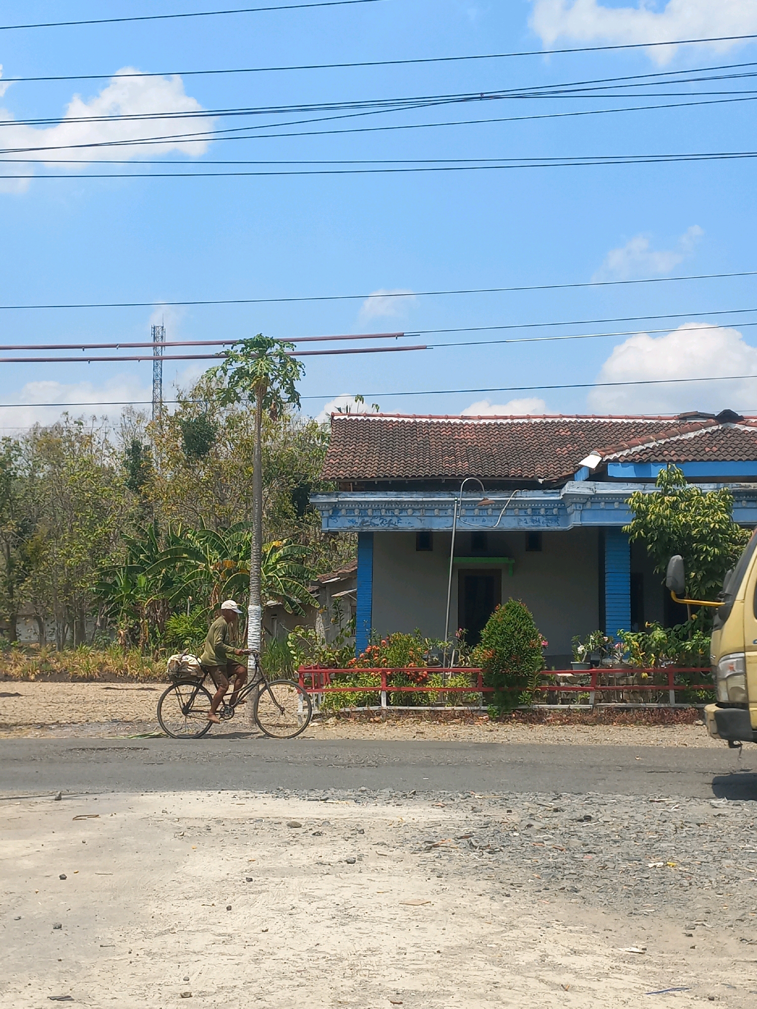 A man is cycling down a paved road in Baturetno, Indonesia. He is wearing a green shirt and a white cap, and is carrying a load of goods on the back of his bicycle. The road is bordered by a row of trees and a house with a blue facade. A yellow truck is parked on the right side of the road. The sky is blue with scattered clouds.
